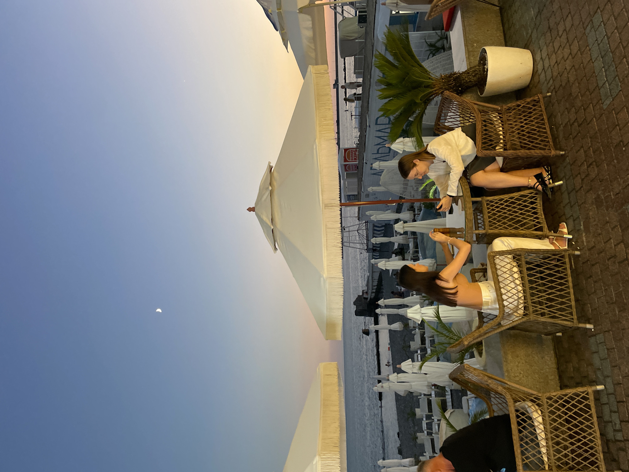 Two young women are seated at a round wicker table outdoors, facing each other. The woman on the left, with long dark hair, is wearing a light-colored top and white pants. She is reaching for something on the table. The woman on the right has blonde hair and is wearing a white long-sleeved shirt and a dark skirt. She is looking down at her phone, which is on the table. They are at a cafe or restaurant with a view of the sea. White beach umbrellas and lounge chairs are visible in the background, suggesting a beach resort setting in Sochi, Russia. The sky is a clear blue with a faint crescent moon visible, indicating it is either early evening or late afternoon. A palm tree in a white pot stands to the right of the table. The ground is paved with bricks. A man in a black shirt is seated at another table to the left, partially visible.