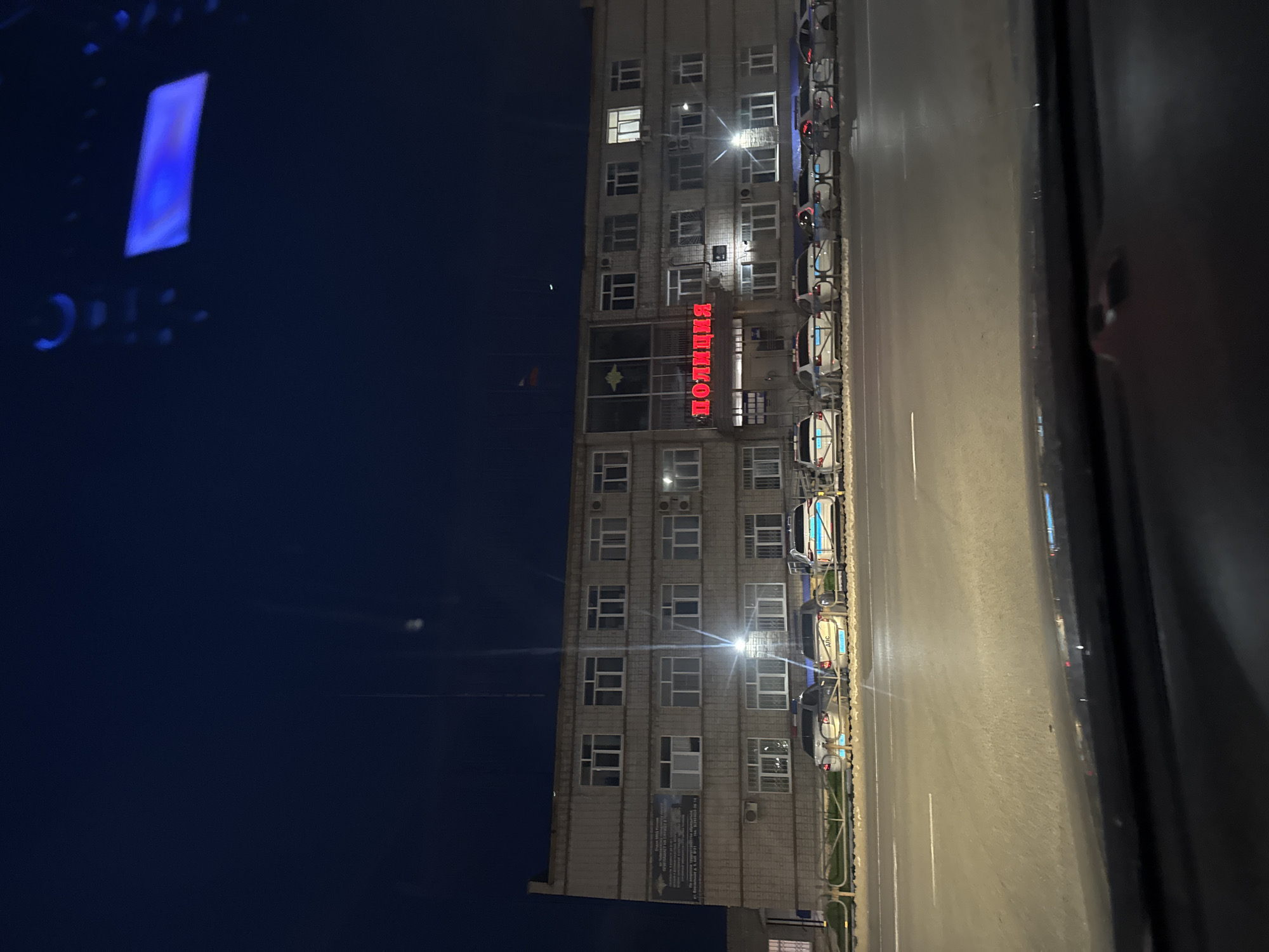 The image shows a building illuminated at night, identified as a police station in Chaikovsky, Russia, based on the Cyrillic script надпись "ПОЛИЦИЯ" displayed in red neon lights. Police vehicles are parked along the street in front of the building. The image appears to be taken from within a vehicle, given the dashboard partially visible at the lower right edge of the frame. The sky is dark, with a faint crescent moon visible.