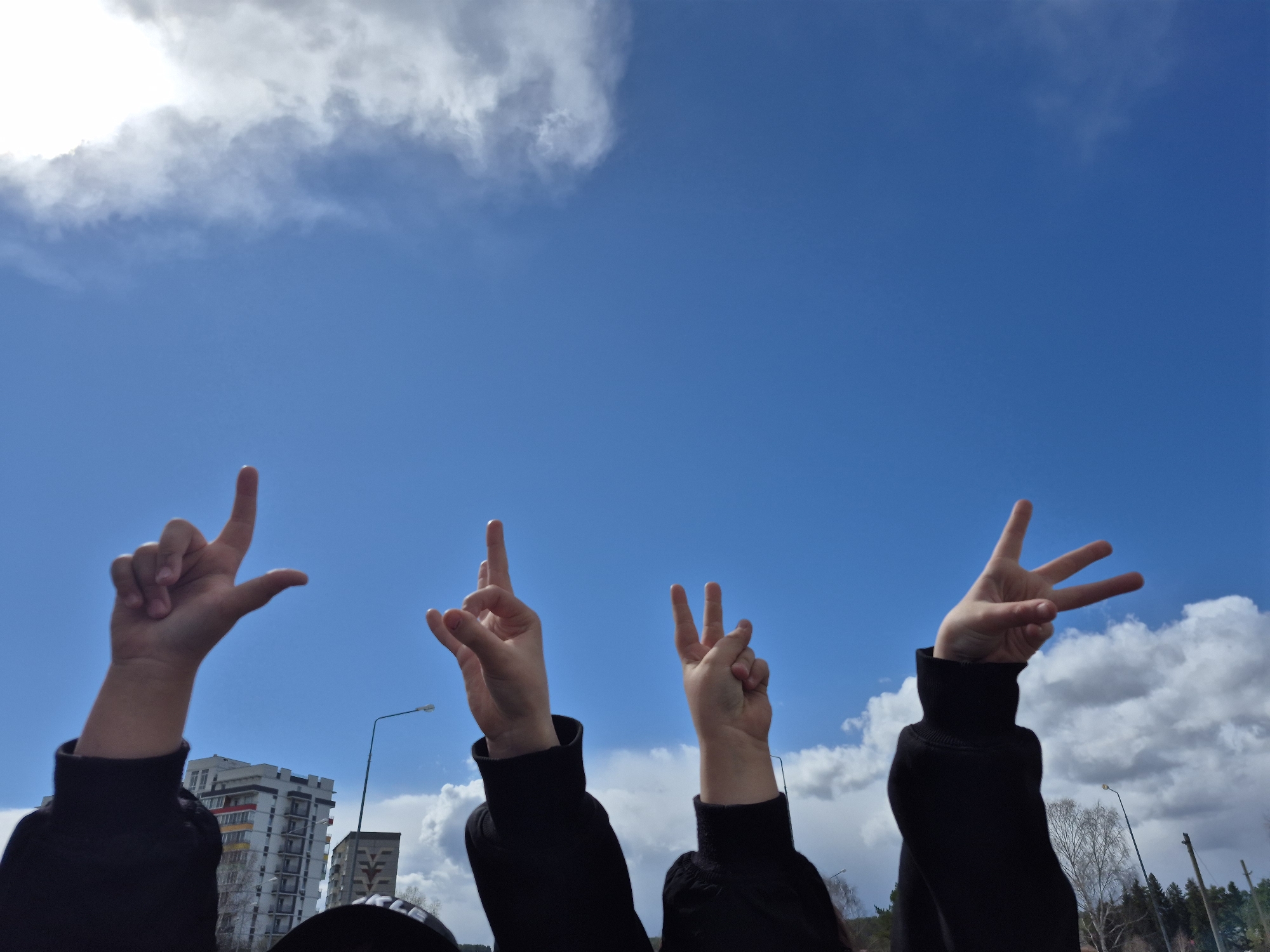 Four hands are raised towards a bright blue sky, positioned across the lower half of the frame, against a backdrop of clouds and distant urban elements. From left to right, the hands perform gestures that spell out the word "LOVE".
The first hand on the left forms the letter 'L' with an extended thumb and index finger. The second hand, positioned slightly to the right, has its index finger pointing upwards and thumb extended horizontally, appearing to represent the 'O' in a stylized manner within the sequence. The third hand displays the 'V' sign with its index and middle fingers extended. Finally, the fourth hand on the right forms the letter 'E' with its index, middle, and ring fingers extended. All individuals are wearing long-sleeved black tops or hoodies.
The sky is predominantly clear blue, with scattered white cumulus clouds, and a large, bright cloud mass is visible towards the top left. The scene suggests a daytime setting with good visibility. In the background, partially visible buildings, a street lamp, and bare trees hint at a late winter or early spring season. To the left, a multi-story residential building with balconies is seen, featuring some colorful panels on one side. Another smaller, darker building with a distinct geometric pattern is visible behind the first hand. A street lamp and dormant trees are visible on the right side.
The location context for this image is Chaikovsky, Russia, which aligns with the architectural style of the buildings in the background. The gestures convey a positive message of love or unity. There is no discernible text visible in the image.