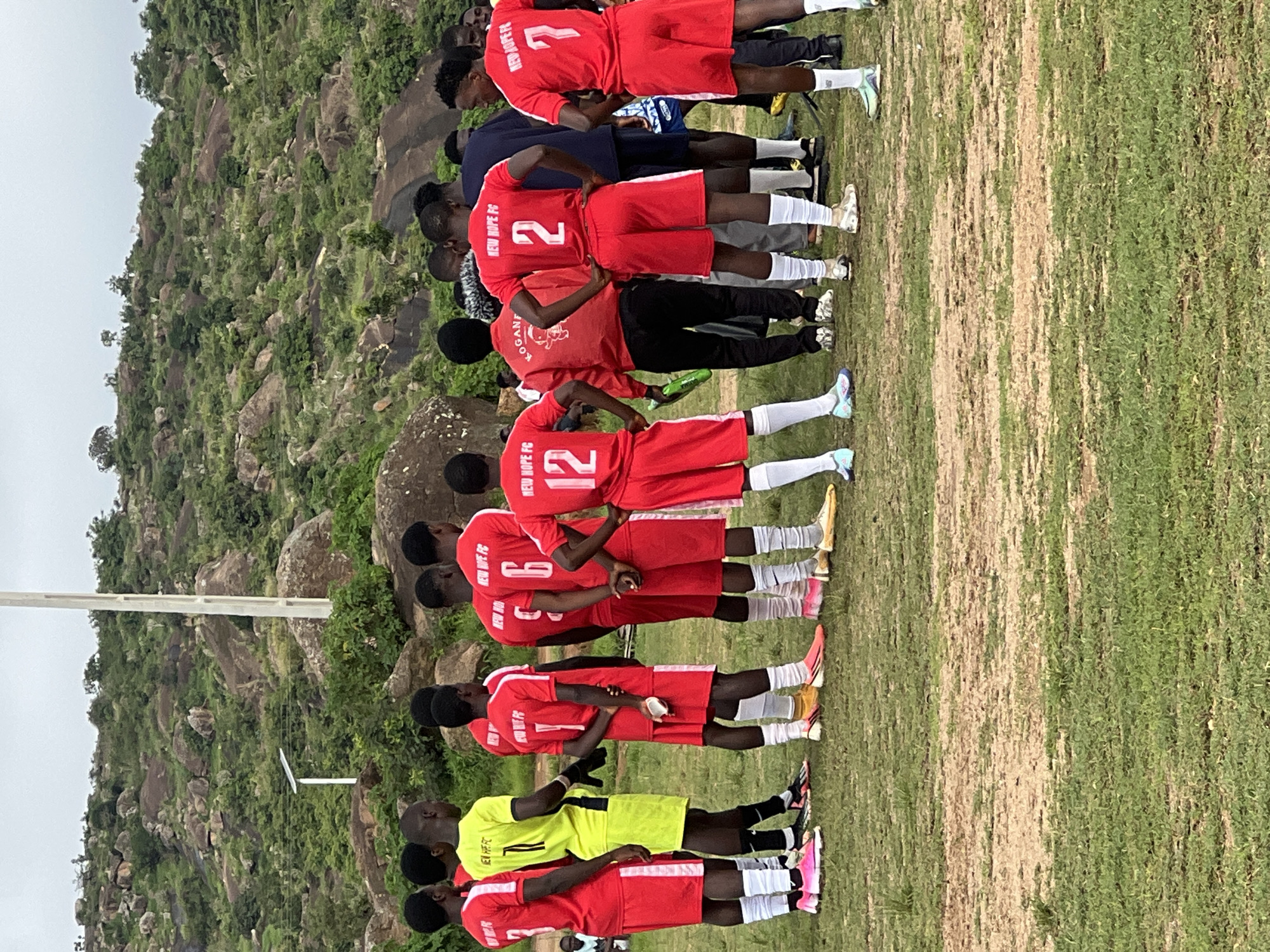 The image captures a group of young men in soccer uniforms, seemingly gathered on a field with a rocky, vegetated hillside in the background. The setting appears to be an outdoor sports facility. The men are mostly wearing red jerseys and shorts, with one player in a bright yellow jersey. Some of the jerseys have numbers and "NEW HOPE FC" printed on them. The ground is a mix of dry, dusty earth and green grass. In the background, a white pole, possibly part of a light or communication tower, stands against the hill. A small solar panel is visible on the hill as well. The overall impression is that of a soccer team, possibly before or after a match, in a rural or semi-rural area near Jos, Nigeria, given the landscape's characteristics.