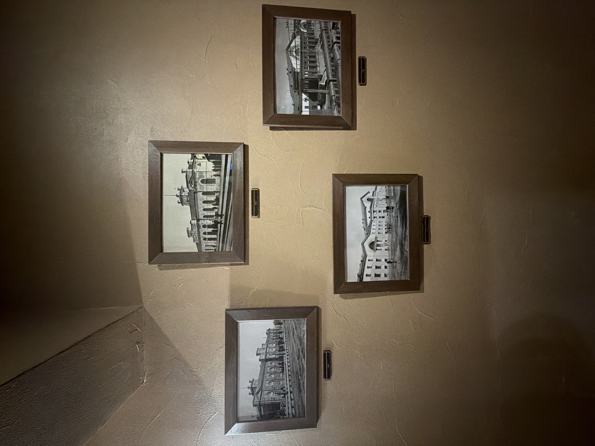 The image shows four framed black and white photographs hanging on a light brown textured wall. The frames are dark brown. The photographs depict architectural structures. The top photograph shows a building interior with staircases and scaffolding. The bottom photograph features a building exterior with multiple windows and architectural details. The photograph on the left shows a building facade and adjacent railroad tracks. The photograph on the right shows a building facade. There is a dark brown rectangle mounted near each frame. A corner of the wall is visible on the lower left. This is in Krasnoyarsk, Russia.