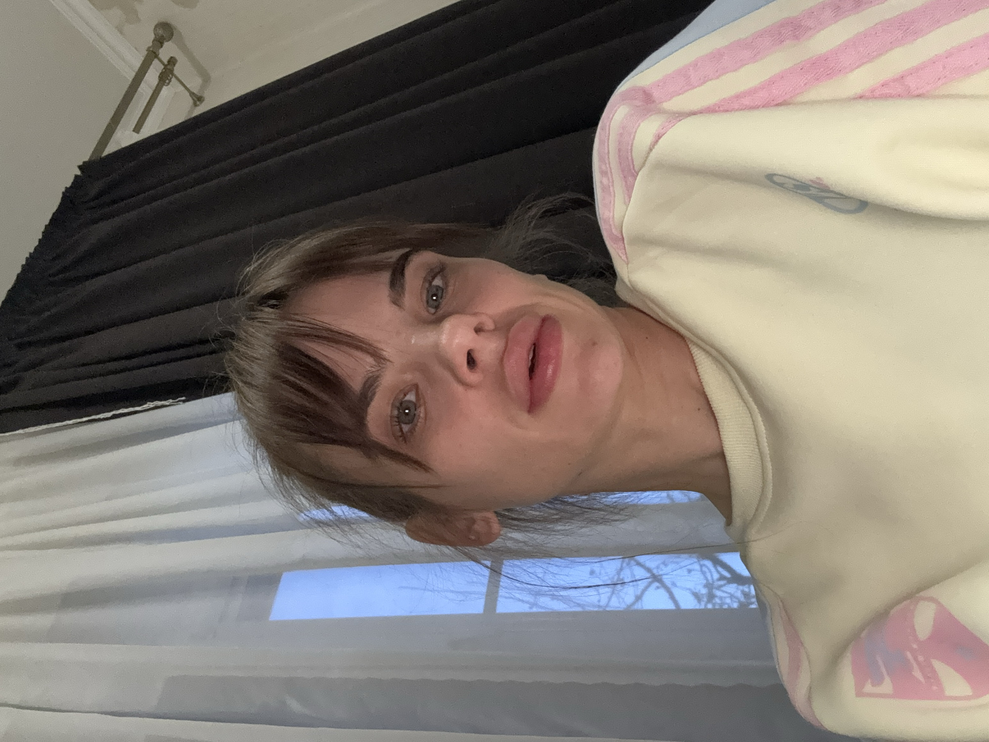 An image shows a young woman's face and upper torso, captured in what appears to be a selfie with a rotated orientation, making her head tilted significantly to her right (viewer's left). She looks directly into the camera with a neutral expression and slightly parted lips. She has light-colored eyes, dark eyebrows, and brown hair with bangs. She is wearing a cream or off-white sweatshirt featuring prominent pink stripes on the shoulder/sleeve area. A faded, light blue or purple graphic logo, possibly stylized initials like "AD", is visible on the front of the sweatshirt.
The background reveals an indoor setting. To the upper left, a section of dark grey or black curtains is drawn, and above them, gold-toned curtain rods are mounted on a white ceiling. To the lower right, a white, possibly sheer curtain or blind covers a window. Through the window, a light blue sky and the silhouettes of bare tree branches are discernible, suggesting daytime during a colder season. The scene takes place in a room located in Rostov-on-Don, Russia.