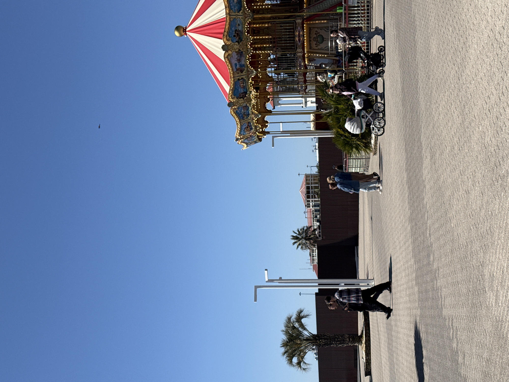 The image captures a vibrant outdoor scene featuring a prominent carousel under a clear blue sky. The carousel is a central focal point, displaying a classic red and white striped canopy, ornate golden embellishments, and numerous decorative lights. Several people are present, enjoying the sunny day.
Near the carousel, a person in a plaid shirt is pushing a stroller, with another individual standing close by. More people can be seen on the carousel itself. In the mid-ground, a person wearing a denim jacket walks across the paved area. Further to the right, another individual in a plaid shirt and dark pants is walking, casting a long, distinct shadow on the ground, indicating bright sunshine.
The ground is paved with light-colored, patterned blocks. The background includes dark brown buildings with white railing accents and several green palm trees. White L-shaped poles, possibly light fixtures or structural elements, are also visible. A small, dark, elongated object can be seen high in the clear blue sky, possibly a bird or distant aircraft. The scene suggests a leisure or entertainment setting, such as an amusement park or public plaza, likely in a warm region given the presence of palm trees. The weather is clear and sunny, and no visible text is discernible.