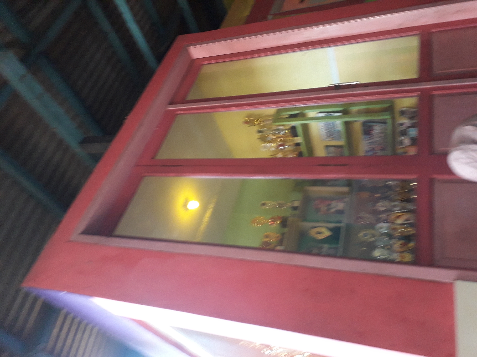 The image shows a close-up, low-angle view of a red-framed display case filled with trophies and medals. The display case has glass doors, through which the items can be seen. On shelves inside the case, numerous trophies of varying sizes and designs are visible, many appearing to be gold or silver in color. Some medals are also displayed. The background within the case is painted a light green and yellow. Above the display case, a corrugated metal roof with blue wooden beams is visible. The overall scene suggests an awards display, possibly in a school or community center. The image is taken in Cankuang, Indonesia, though the exact location within the city is not specified.