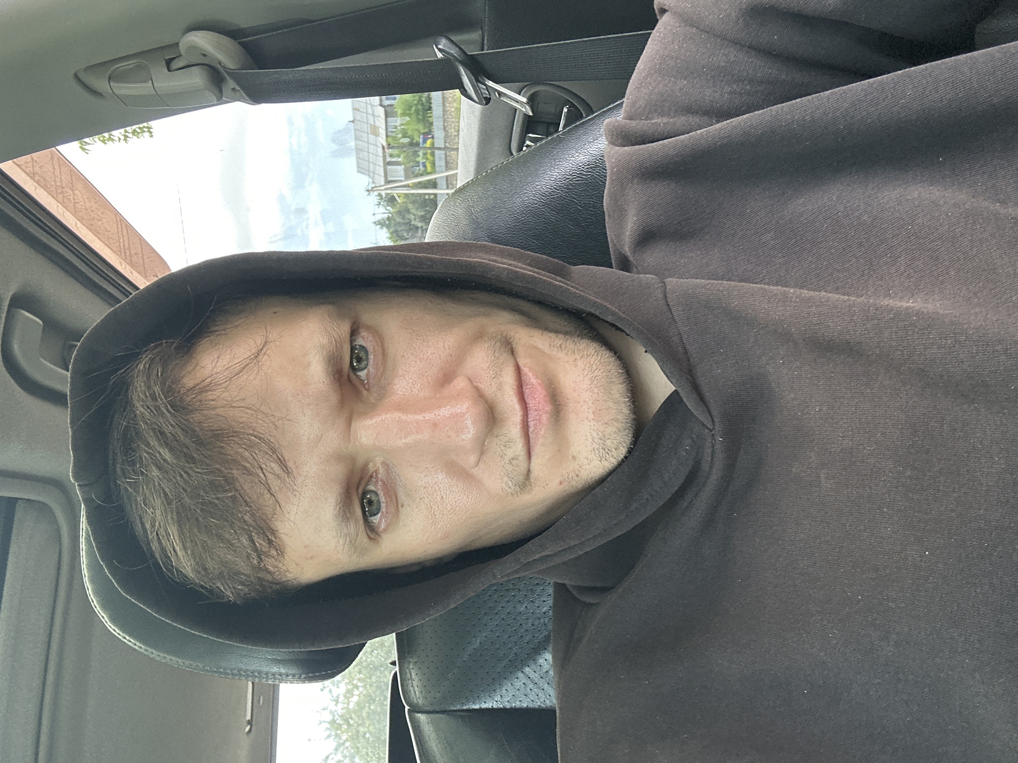 A man with light brown hair and green eyes, wearing a dark brown hooded sweatshirt, is seated in the driver's seat of a car. He is looking directly at the camera. The interior of the car, including a black leather seat and a seatbelt, is visible. Outside the car, a cloudy sky and residential buildings can be seen. The image appears to have been taken outdoors in the city of Makiv, Ukraine.