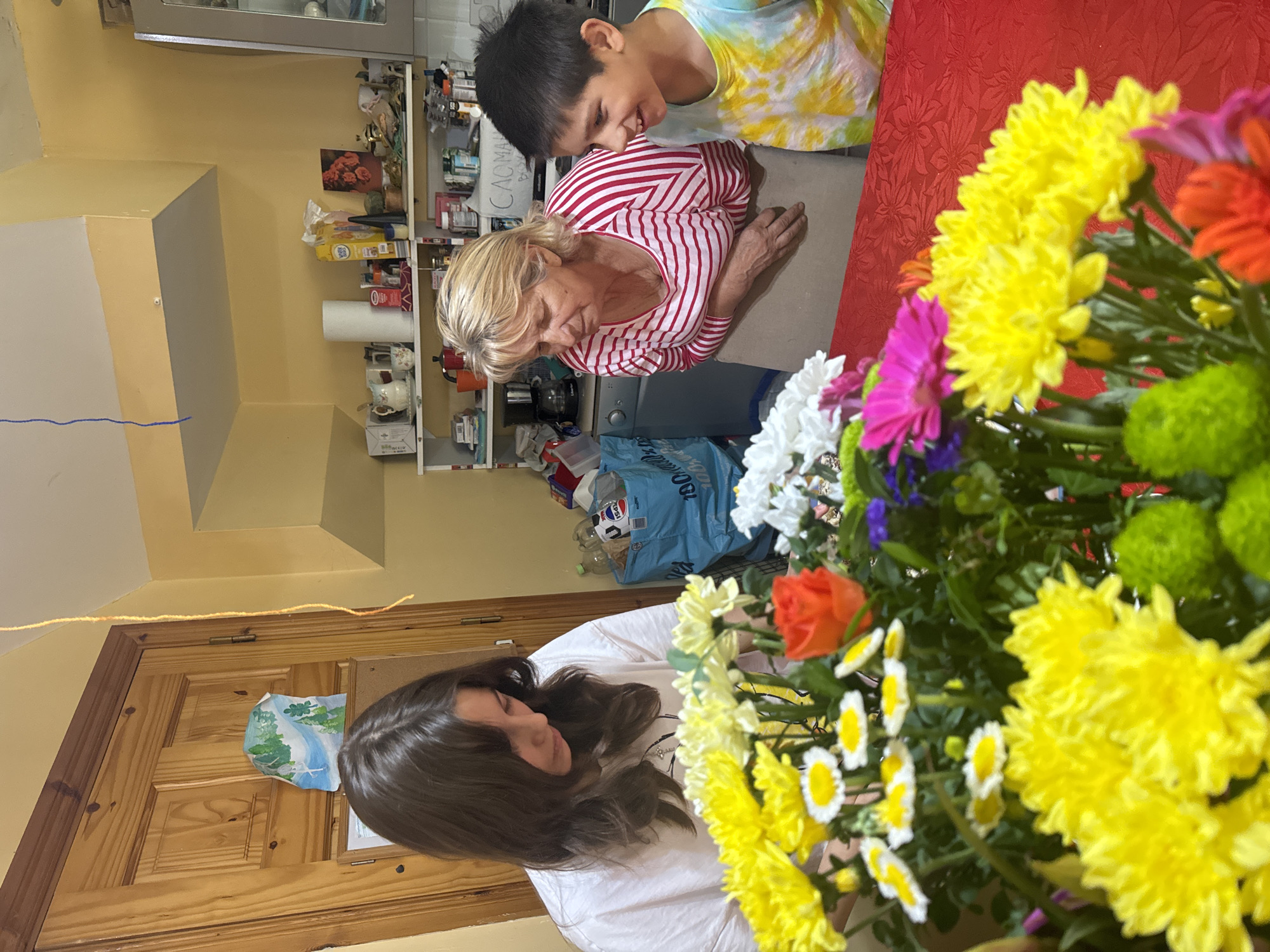 An indoor scene depicts three people in what appears to be a kitchen area, with a focus on a colorful floral arrangement. A young female with long, dark brown hair, wearing a white t-shirt, is positioned on the left, looking down at the flowers. To her right, an older woman with blonde hair, dressed in a red and white striped long-sleeved shirt, leans forward, observing something with a slight smile. Next to her, a young male with dark hair, wearing a tie-dye t-shirt, beams with a wide smile. The floral arrangement, predominantly featuring yellow, orange, white, and pink flowers, is centrally located on a red tablecloth. In the background, kitchen cabinetry, shelves stocked with various items, and a wooden door are visible. The lighting is warm, suggesting an indoor setting. The scene does not contain enough identifiable landmarks to determine a specific location within Waterford, Ireland.