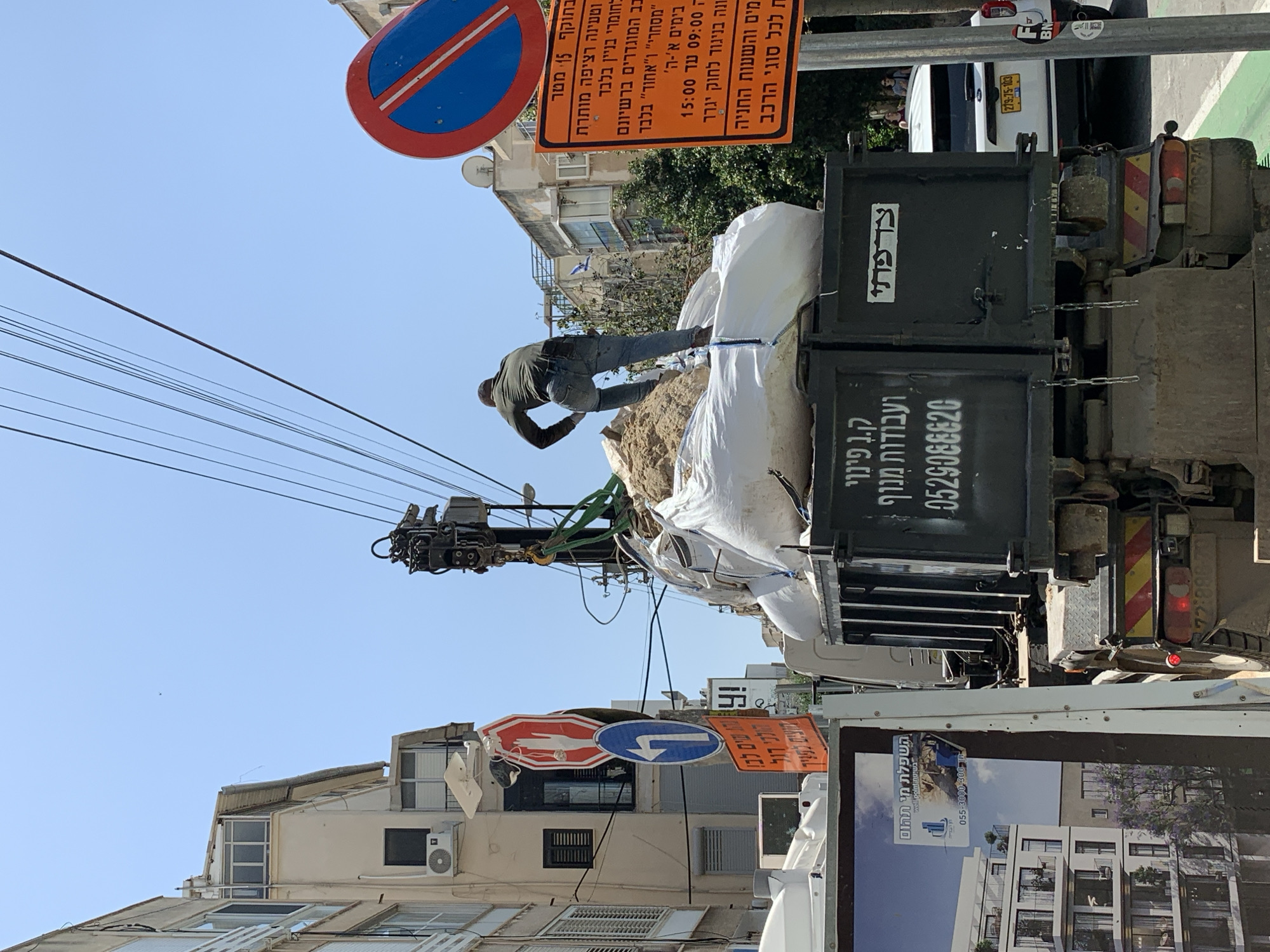 A man is atop a truck bed with large white bags in Tel Aviv, Israel. He is wearing dark clothing. The truck has a crane arm. Numerous street signs with Hebrew text are visible, including a No Parking sign. Power lines extend across the sky. A building with an air conditioning unit is nearby. A white car with a yellow license plate is parked nearby. There is a construction project. The truck has Hebrew words "Pina" and a phone number on it.