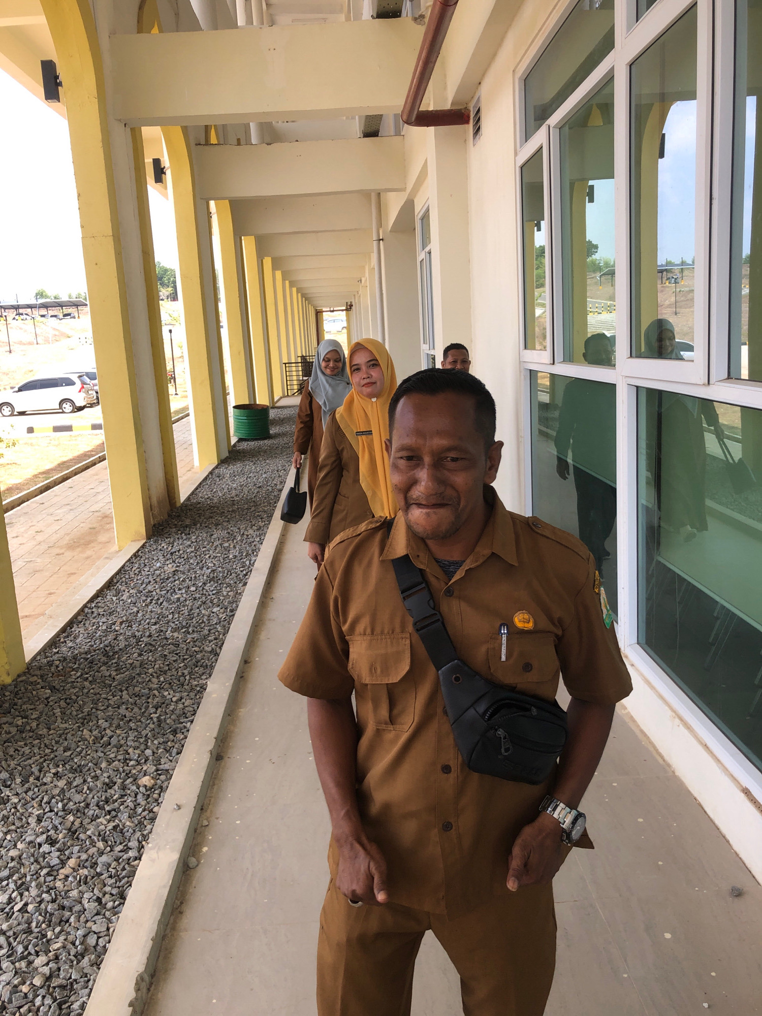 In Reuleut Timu, Indonesia, a group of four individuals, three women and one man, are captured in a medium shot. The man in the foreground, wearing a brown uniform, faces the camera with a neutral expression. A black fanny pack is slung across his chest, and a watch adorns his left wrist. Behind him, three women in matching brown uniforms and headscarves walk in a line. The woman in the yellow headscarf is closest to the man, followed by two women in light blue headscarves. The background reveals a covered walkway with yellow pillars and windows reflecting the sky and surrounding buildings. Outside the walkway, a gravel-covered area lines the path, with a white car visible in the distance. The overall scene suggests an outdoor setting, possibly at an educational institution or government building.