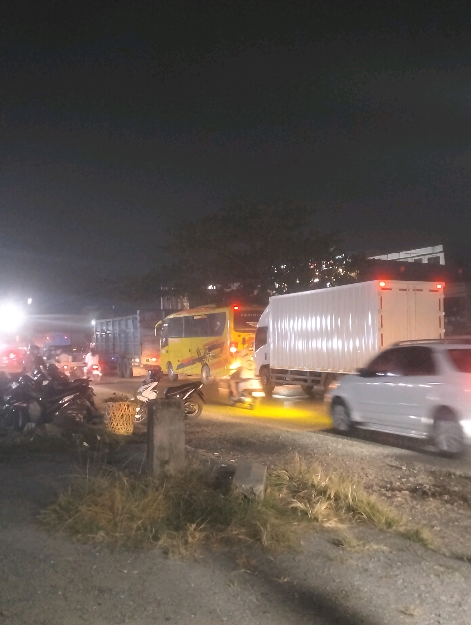 At night in Tambusai Batang Dui, Indonesia, a traffic jam is depicted on a paved road. The scene is illuminated by vehicle headlights and streetlights, casting long shadows. Multiple vehicles are present, including a large white truck with a cargo container, a yellow bus with "PARIWISATA" branding, a blue truck, and several motorcycles parked on the left side of the road. A white car is in motion on the right side of the frame. Vegetation, including grass and a woven basket, are visible along the side of the road. The sky is dark and appears overcast.