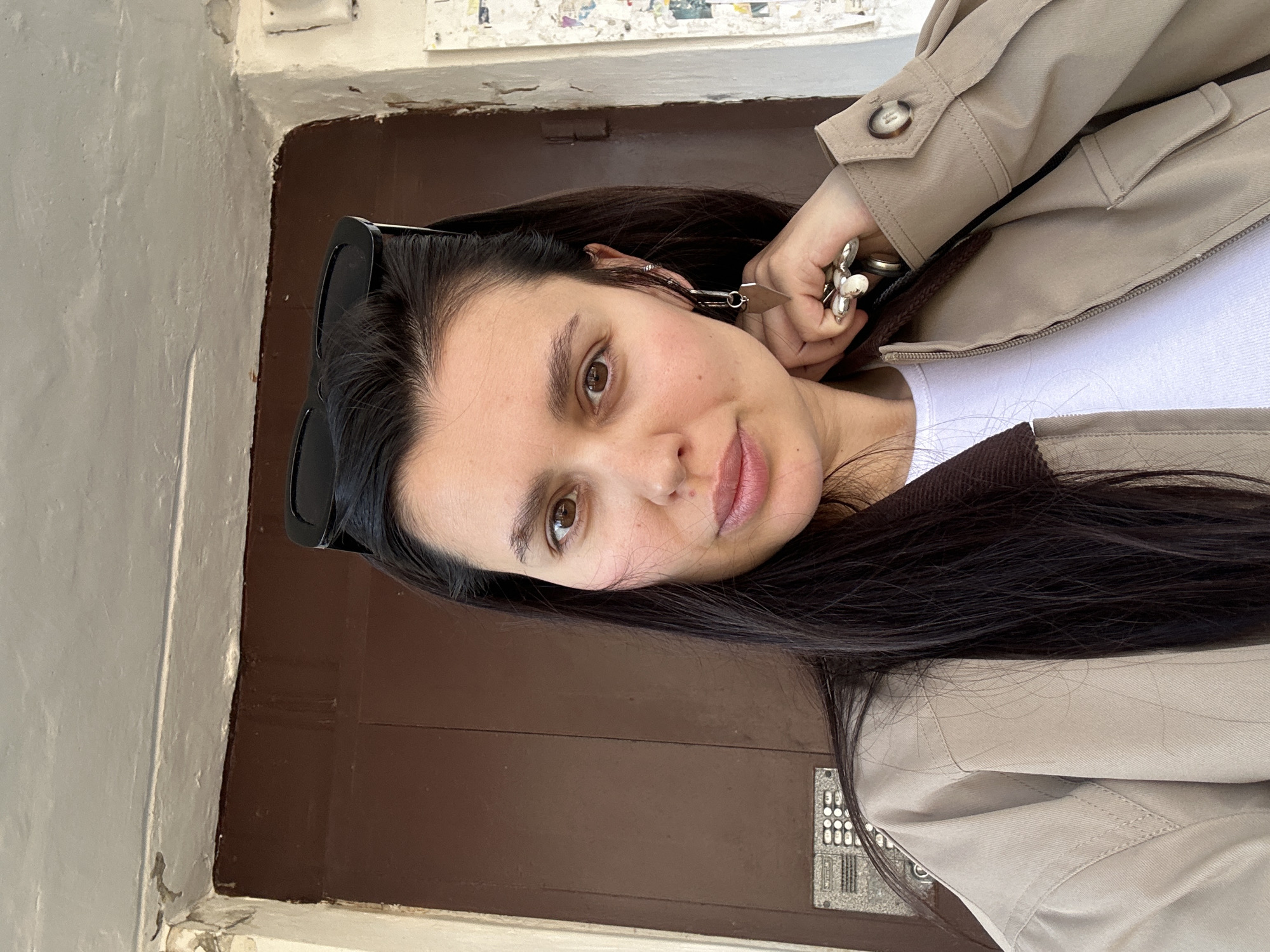 A young woman poses for a selfie or portrait. She has dark brown hair and brown eyes. She is wearing sunglasses and a beige jacket. The background appears to be a brown doorway with a weathered white frame. A metal intercom panel is visible on the door. Based on available geographical information, the photo was taken in Chaikovsky, Russia.