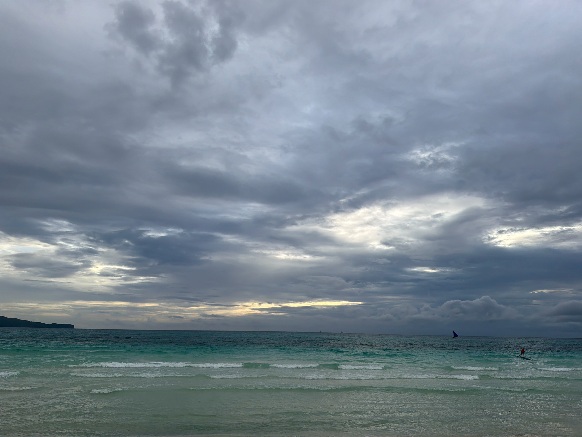 This image captures a scenic view of a tropical beach, likely in Boracay, Malay, Philippines, under a dramatic, overcast sky. The ocean exhibits varying shades of turquoise and deep blue, with gentle waves lapping at the shore. In the distance, a sailboat with a purple sail is visible, and closer to the foreground, a person is standing on a paddleboard, facing the ocean. The horizon is marked by a faint line of land on the left side, appearing as a dark silhouette against the bright sky. The sky itself is filled with thick, grey clouds, punctuated by patches of lighter sky, suggesting either an impending storm or the lingering aftermath of one. The overall atmosphere is moody and serene, characteristic of a cloudy day at a tropical seaside.