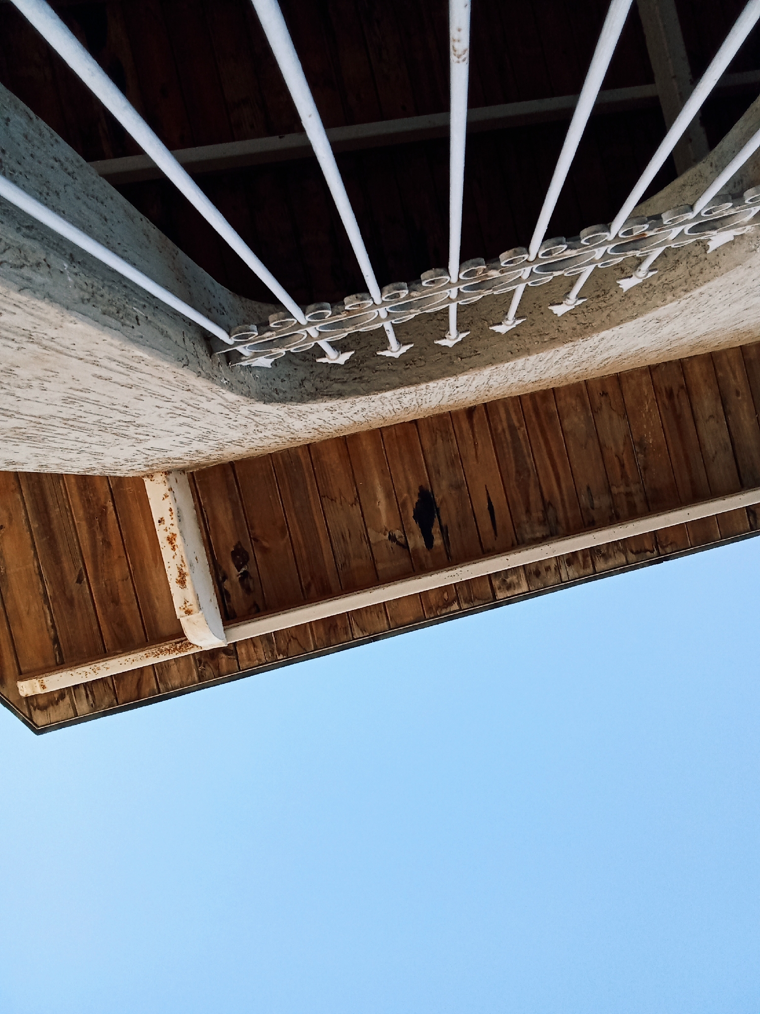 A low-angle shot captures an overhanging structure against a clear blue sky, likely an architectural detail in the city of Juan Griego, Venezuela. The foreground is dominated by the sky, which fades into the edge of the building. The overhang features a wooden paneled ceiling with visible imperfections and discoloration, suggesting weathering or age. A white metal frame supports the wooden structure, showing signs of rust. An ornamental white metal grille is affixed above the wooden paneling, creating a decorative effect.