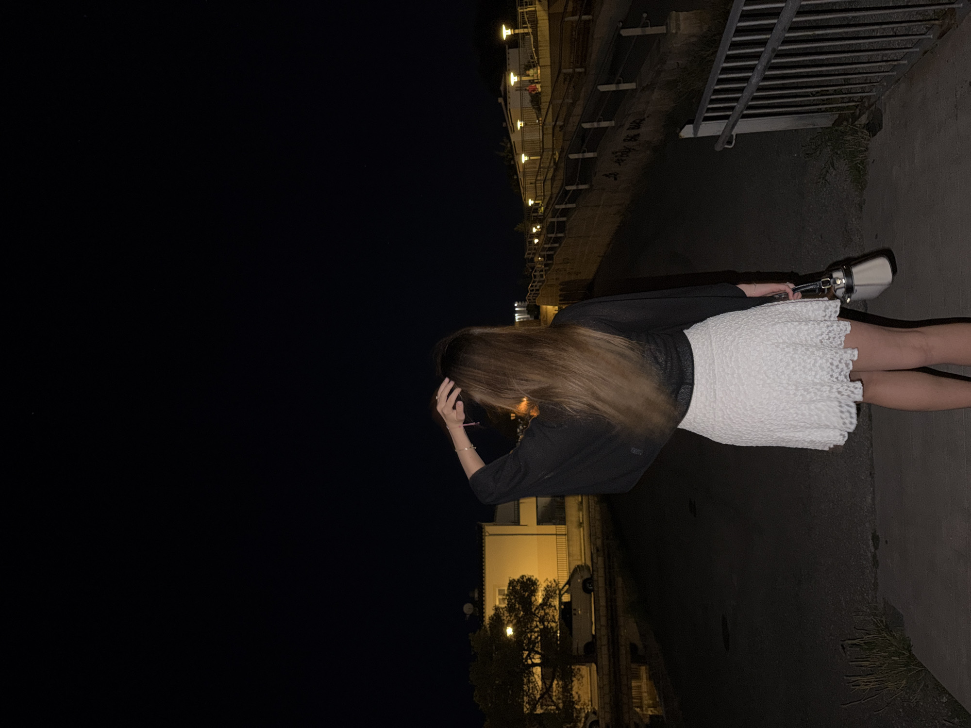 A nighttime outdoor photograph shows a woman with long brown hair standing on a concrete sidewalk, wearing a short white lace dress and a dark jacket. She is facing away from the camera and appears to be adjusting her hair with her right hand. A white purse with black hardware is held in her left hand. The sidewalk is adjacent to a dark, paved area, possibly a street or walkway. In the background, buildings with illuminated windows and streetlights are visible, suggesting an urban setting. Based on the distinctive architectural style and street lighting, this photo was taken in Alcúdia, Spain. A metal railing is situated to the right of the woman. The sky is black and empty.