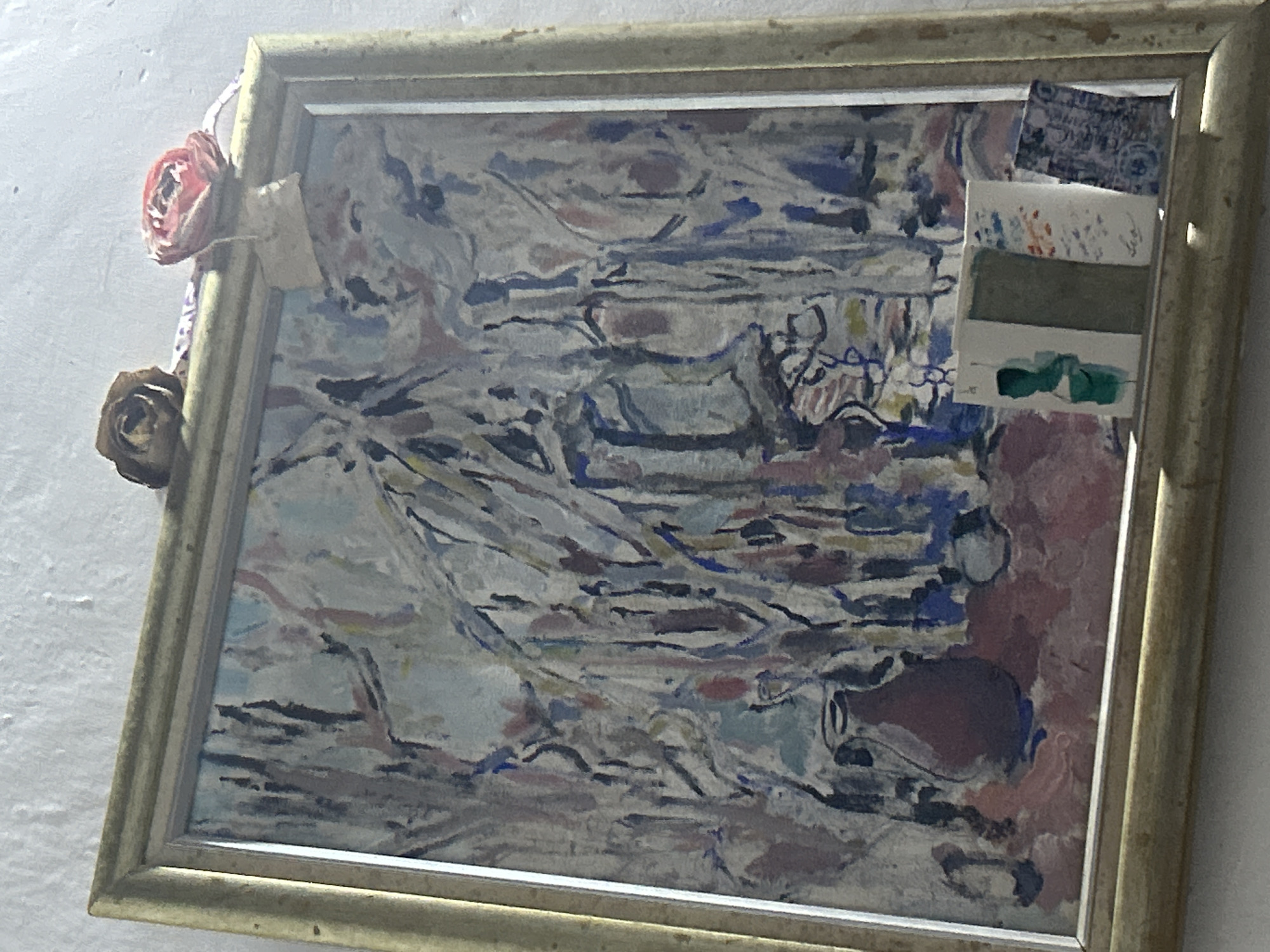 The image shows a framed abstract painting with a textured, white wall behind it. The painting is gold-framed and appears to be hanging on the wall. Resting on top of the frame are two dried roses, one brown and one reddish-pink, attached with a ribbon. Below the painting, on a shelf or ledge, are three small items: a white card with a green silhouette, a white card with abstract colored marks and what appears to be a signature, and a dark blue patterned card. The painting itself is rendered in a loose, impressionistic style with a palette of blues, grays, whites, reds, and yellows, depicting what could be interpreted as a still life with a dark vase on a pink surface, and abstract forms in the background. The overall impression is of a domestic interior setting, likely a home. The question about the location in Santa Ana, Venezuela, cannot be answered from the provided image.