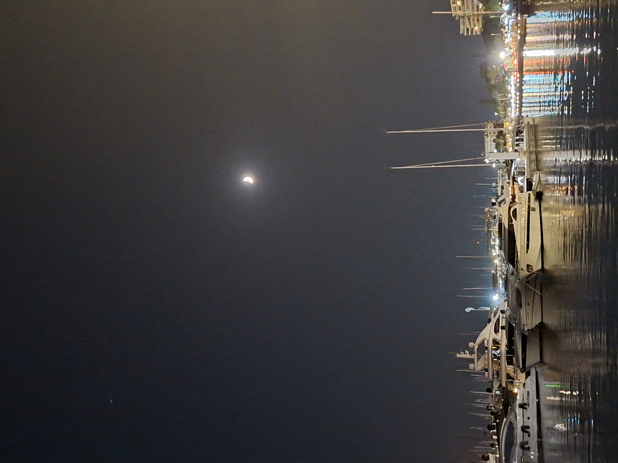 The image depicts a nighttime scene at a marina in Alicante, Spain. The sky is dark with a crescent moon visible in the upper right quadrant, accompanied by a faint star or satellite in the upper left quadrant. The foreground shows the calm water of the marina, reflecting the lights from the city and the docked boats.
Numerous yachts and boats are moored in the marina, showcasing various sizes and designs. Their lights are illuminated, contributing to the ambiance of the scene. In the background, the city lights of Alicante create a bright, colorful glow, with buildings and possibly palm trees visible. The overall impression is one of a peaceful, urban waterfront at night.