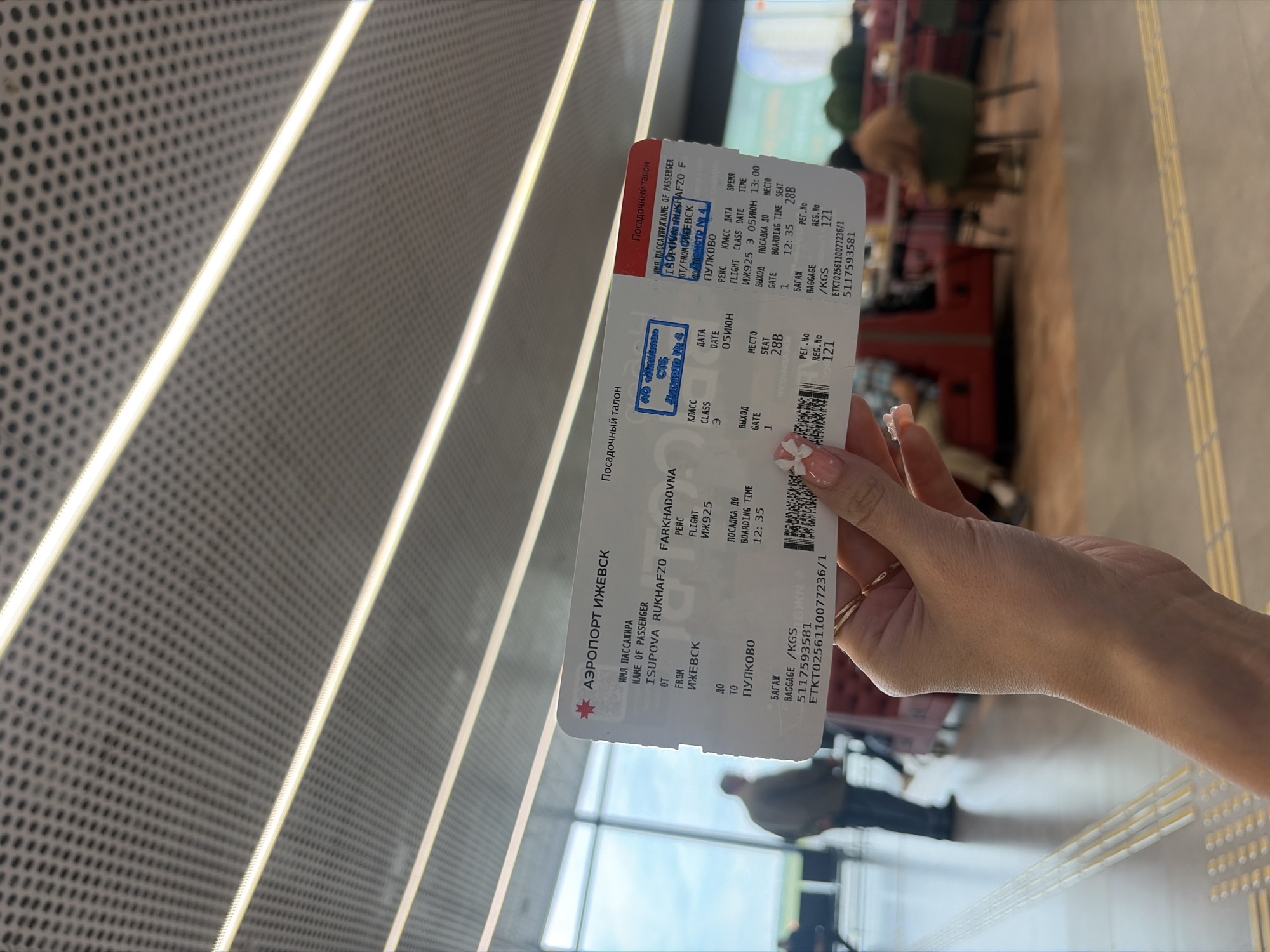 A close-up shot captures a hand holding a boarding pass in what appears to be a modern airport terminal. The hand, adorned with light-colored manicured nails, holds the white and red boarding pass diagonally across the frame.
The boarding pass clearly displays details in Russian and English. It identifies the passenger as ISUPOVA RUKHAFZO FARKHADOVNA, flying from "ИЖЕВСК" (Izhevsk) to "ПУЛКОВО" (Pulkovo, likely St. Petersburg). The flight number is ИЖ925 (IZh925), scheduled for 05ИЮН (June 5th) at 13:00, with boarding time at 12:35. The passenger's seat is 28B, and the gate is 1. Visible stamps on the pass include "АО «ИЖАВИА»" (JSC "Izhavia") and "Досмотр № 4" (Security check № 4), confirming the origin airport and security clearance. Baggage information shows "121" for number or weight.
The background is softly blurred, revealing an indoor setting with contemporary airport architecture. A textured wall or ceiling with a pattern of small holes is visible, intersected by bright, parallel light strips. The floor is light-colored, featuring yellow tactile paving strips on the right. Blurred figures of other people, possibly passengers or airport personnel, and seating areas with tables and chairs can be discerned in the distance, suggesting a waiting lounge or boarding area. The overall scene depicts a traveler's moment within a bustling airport environment at midday.