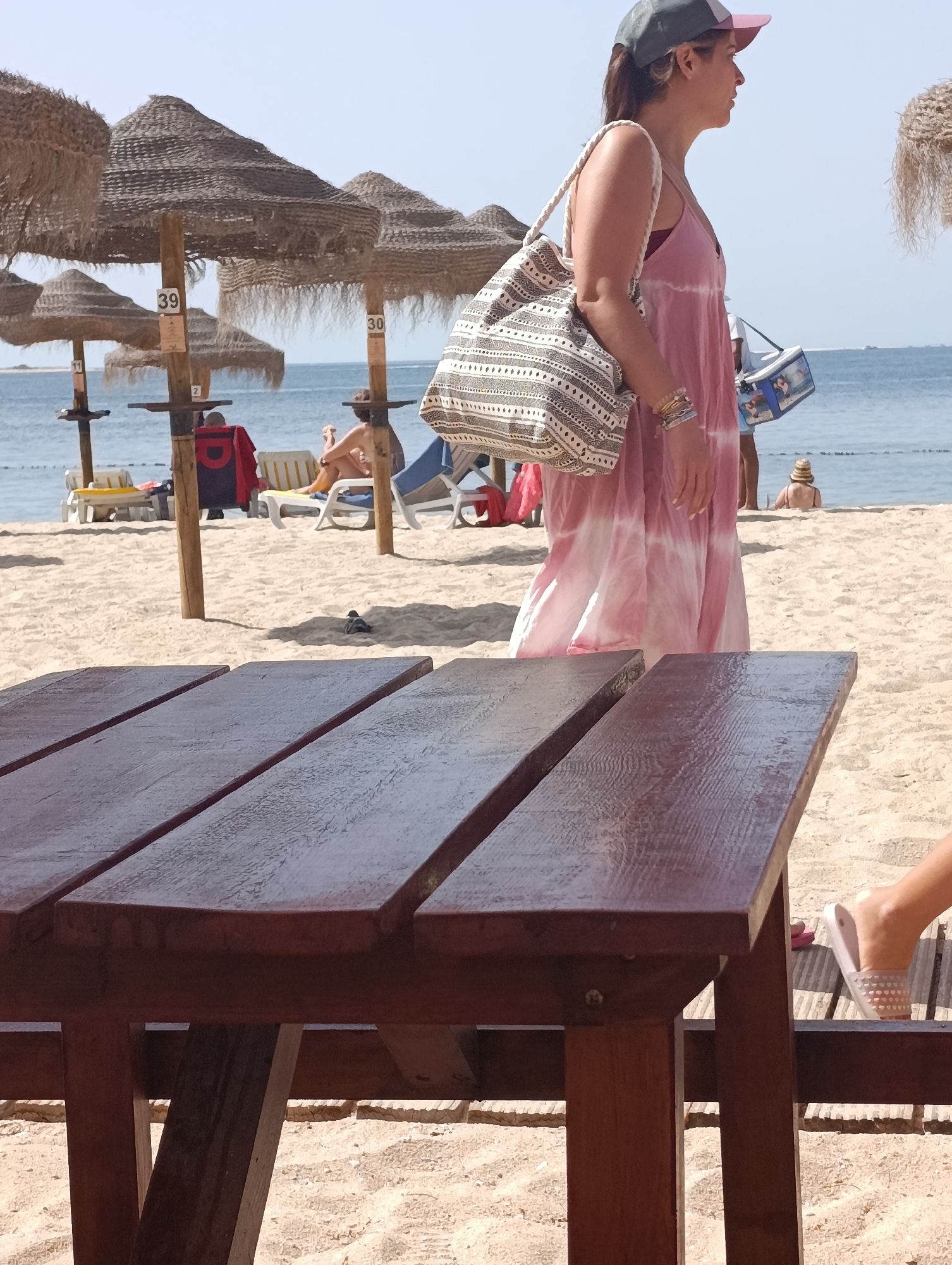 The image depicts a sandy beach scene. In the foreground, a dark wooden picnic table occupies the lower portion of the frame. Behind the table, a woman wearing a pink tie-dye dress and a baseball cap walks away from the camera, carrying a patterned tote bag and a small cooler bag. She is adorned with several bracelets. Further back, beach umbrellas with straw coverings are visible, some with numbers "39" and "30" attached to their poles. Beach chairs are scattered on the sand, with several individuals seated or lounging. The sea forms the background, with a hazy horizon. The overall atmosphere suggests a sunny day at the beach. It is not possible to determine the specific location in Setúbal, Portugal from the image alone.