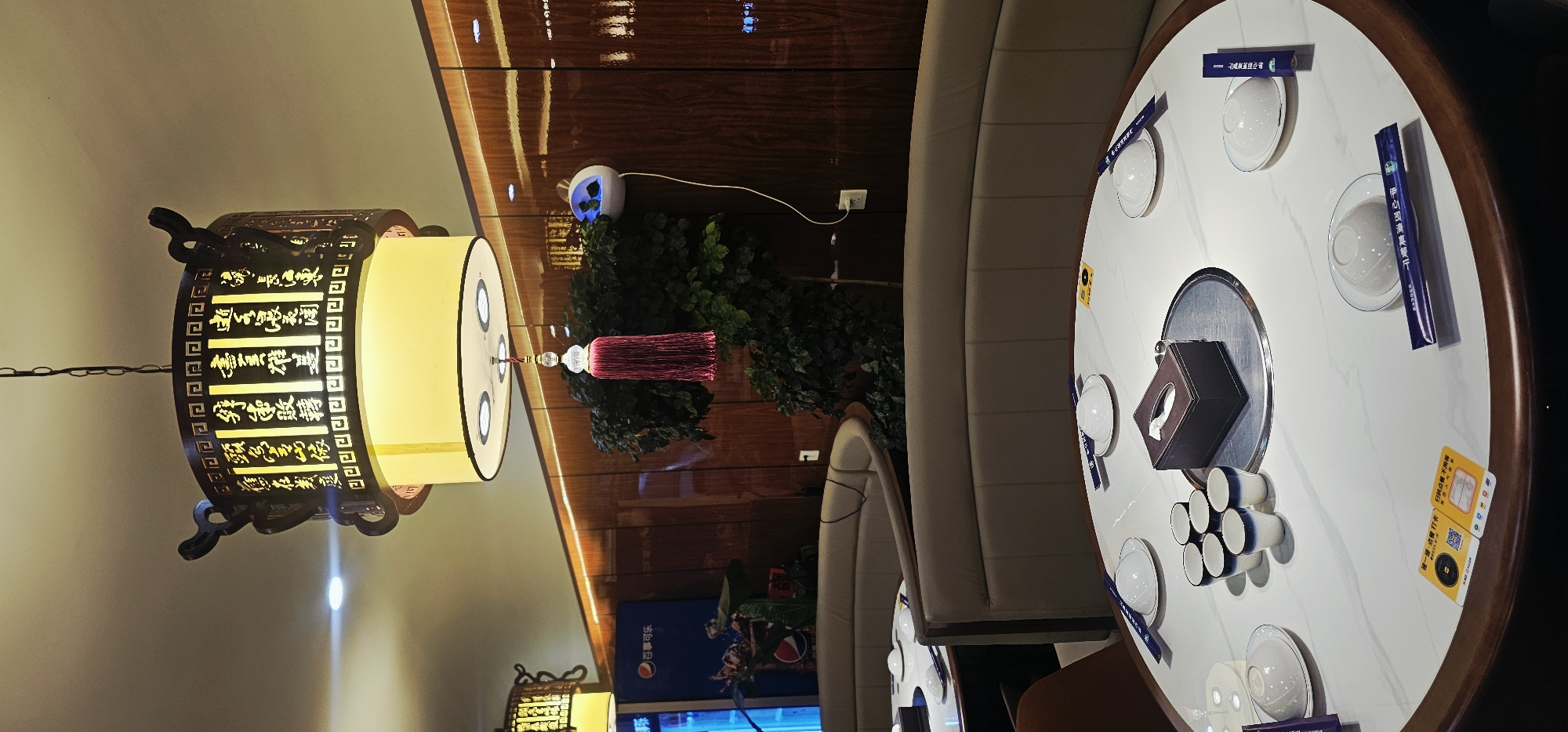 This is a photograph of the interior of a restaurant in Yuzhong District, China. The scene is set at a round marble table, which is set for dining with white bowls, plates, and chopsticks. A dark brown tissue box sits at the center of the table, along with a silver lazy Susan. Two QR code stickers are visible on the table, one yellow and one orange. The table is surrounded by a curved, beige upholstered booth. In the background, a large green plant is visible, and further back, a wooden wall with embedded lights is present. A distinctive overhead light fixture with intricate carvings and a red tassel hangs above the table. A portion of a Pepsi refrigerator is visible to the left of the plant.