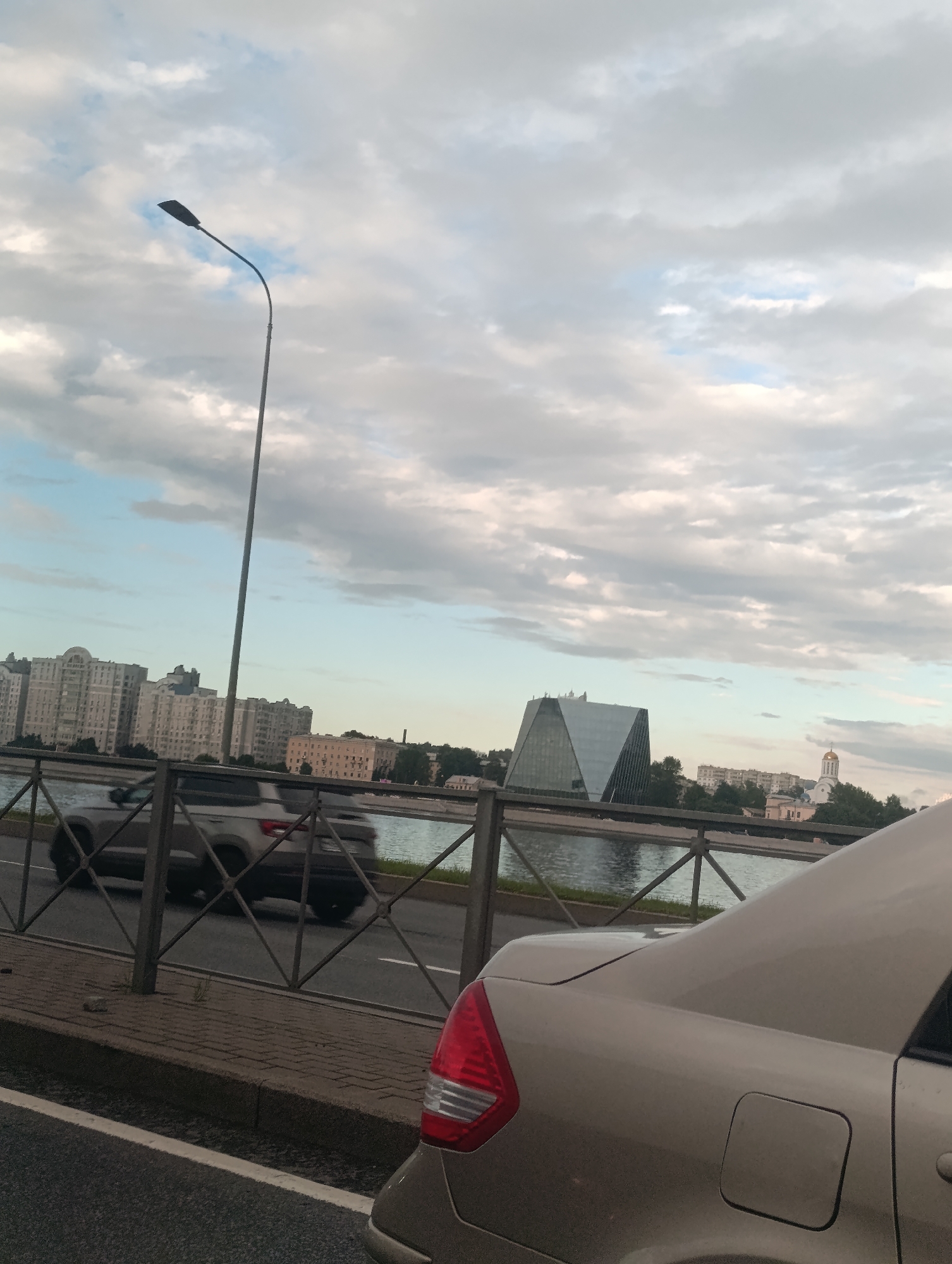 This image captures a view of the Neva River in Saint Petersburg, Russia, under a cloudy sky, likely during the late afternoon or early evening. The foreground is dominated by the rear of a light-colored car, its taillight prominently visible. Beyond the car, a metal fence separates a sidewalk from the Neva River.
Across the river, a cityscape unfolds with several notable structures. On the left, large residential buildings with many windows line the waterfront. In the center, a distinctive modern building with a geometric, angular design stands out. To the right, a white church with a golden dome and a cross is visible, characteristic of Russian Orthodox architecture. Trees dot the landscape between the buildings, adding greenery to the urban scene.
Cars are seen driving on a road parallel to the river, indicating ongoing traffic. The weather appears overcast with patches of lighter sky, suggesting a potentially mixed or approaching storm condition. The overall mood is that of an urban scene with elements of both modern and historical architecture, set against a dynamic sky and water. There is no visible text in the image.