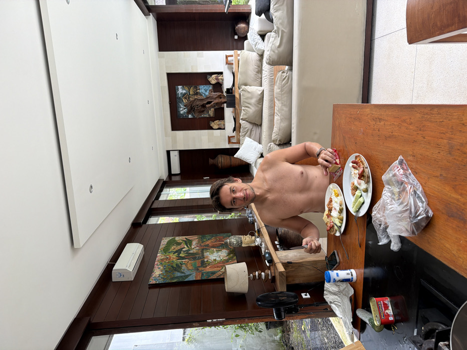 An adult male is seated shirtless at a wooden dining table in a residence located in Ungasan, Indonesia. He holds a knife in his right hand over a white oval plate containing fried eggs, sausages, and cucumber slices. His left hand is positioned near a red-wrapped item on the table, adjacent to a second identical plate of food. A smartphone, a salt/pepper shaker, and a crumpled white paper are also on the table.
To the left of the table, a black electric fan stands on the floor. On a dark surface in the immediate foreground, a red "PRONAS" brand tin can and a clear plastic bag containing unknown contents are present.
The room features dark wooden paneled walls and a light-tiled floor. Along the left wall, large glass sliding doors reveal an exterior view of overgrown greenery and a concrete wall. Above a wooden console table with a lamp and a Buddha head statue, a large, colorful painting hangs. A white air conditioning unit is mounted high on this wall.
In the background, a light-colored sectional sofa with cushions is arranged in a living area. Beyond the sofa, another artistic display is visible on a light-colored wall, featuring a large wooden sculpture in front of a framed landscape painting. Two small, light stools are positioned below this artwork. A second air conditioning unit is mounted high on the back wall. The ceiling is white with recessed lighting and a raised rectangular panel. Large windows or doors are also visible on the right side of the room.