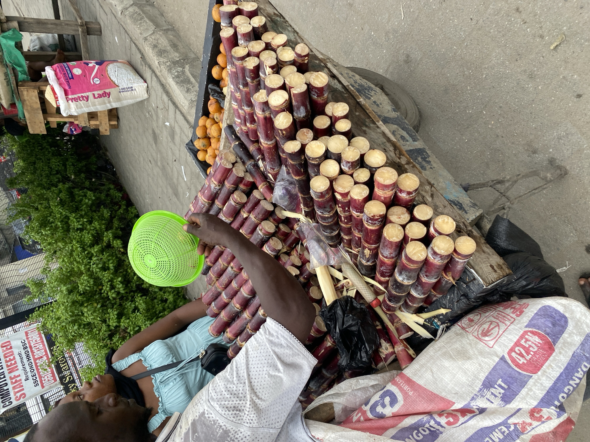 The image depicts a street scene in Lagos, Nigeria, featuring a sugarcane vendor. The vendor, a dark-skinned man wearing a white patterned shirt, is holding a green plastic colander, possibly for rinsing the sugarcane. The cart is stacked with cut sugarcane stalks, arranged in a pyramid formation. A smaller quantity of round, orange citrus fruits is visible alongside the sugarcane. A woman in a light blue dress stands beside the vendor. A cement bag with "Dangote Cement 42.5N" printed on it is near the sugarcane. In the background, partially visible, is a shop window, a "Pretty Lady" brand rice bag on a wooden pallet, and a bush. The environment appears to be a busy street, with other unidentified items and people subtly present. The perspective is from above, looking down at the vendor and his merchandise.