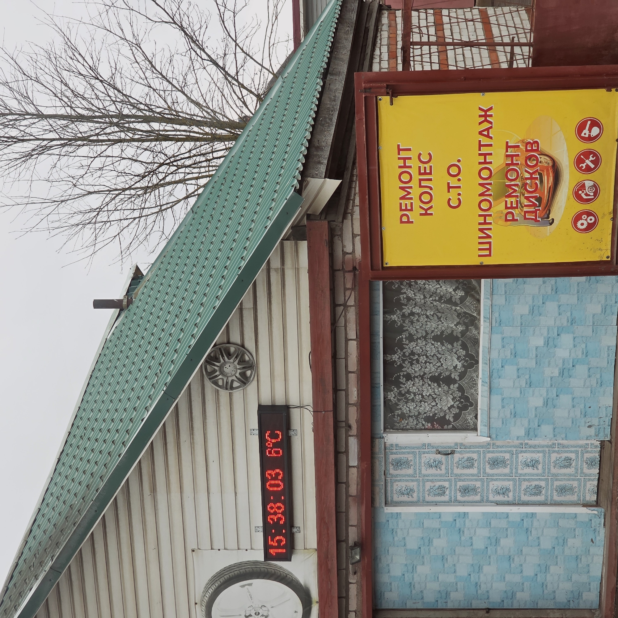 The image shows the exterior of a building, seemingly a tire repair shop ("Ремонт Колёс"). The building has a light-colored, corrugated metal facade and a green corrugated metal roof. A digital display shows the time as 15:38:03 and the temperature as 6 degrees Celsius. A decorative car wheel is mounted on the facade, both near the top and bottom of the building. There is a large yellow sign advertising tire services in Russian. The building shows signs of age and disrepair, with mismatched blue tiles decorating the lower part of the wall and a patterned window above. The location, given the advertisement in Russian and the city specified, is likely Volosovo, Russia. Bare tree branches are visible in the upper left corner of the image.