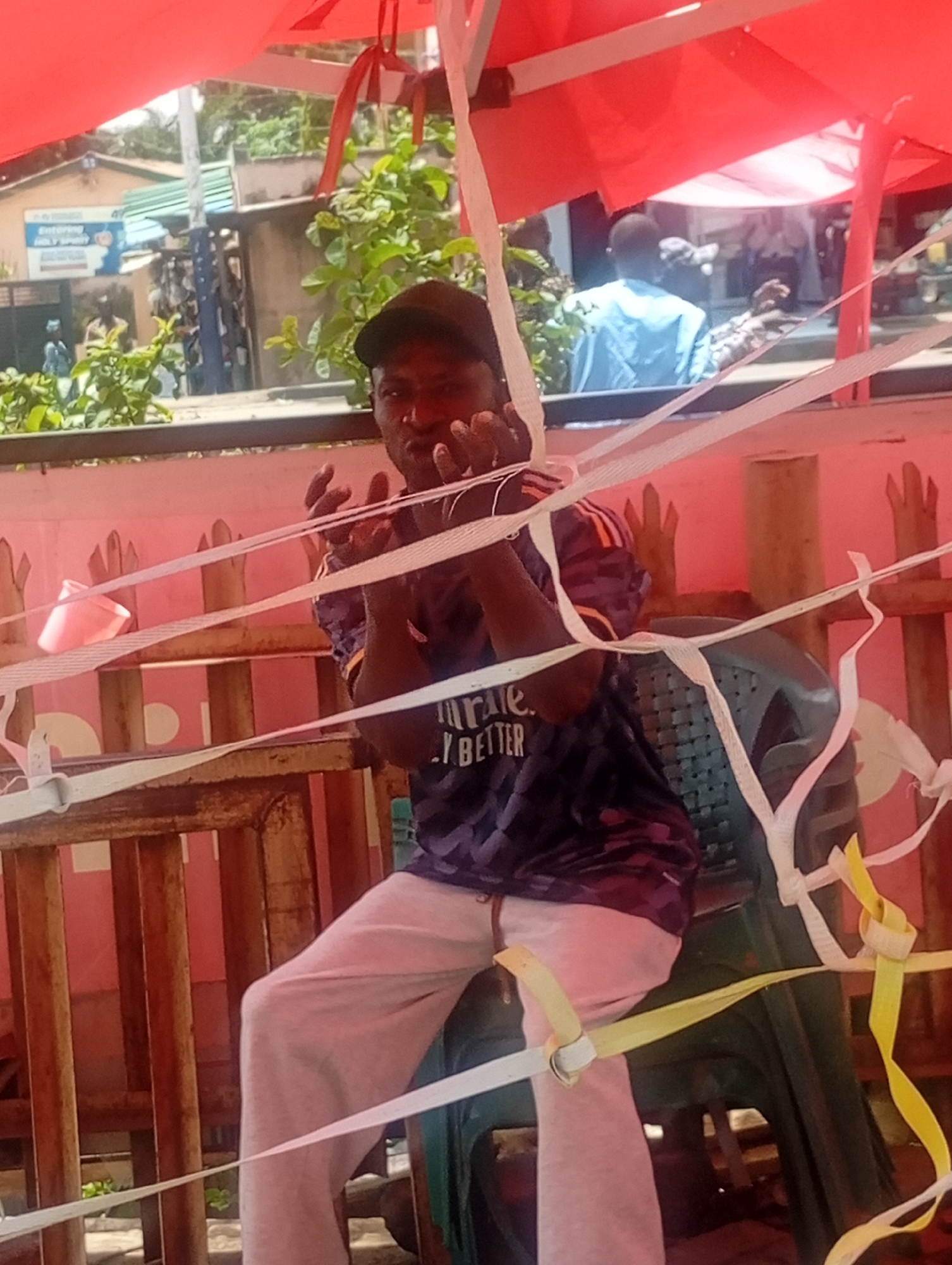 A man wearing a dark baseball cap, a purple and black patterned shirt with white text, and light pink sweatpants sits on a green plastic chair within a temporary structure in Maiburiji, Nigeria. The structure is defined by a red canopy overhead and decorated with white and yellow ribbons. The man is holding his hands up in front of him, palms facing outward. The background includes a pink wall, greenery, and buildings with signs. Other individuals are visible in the blurred background.