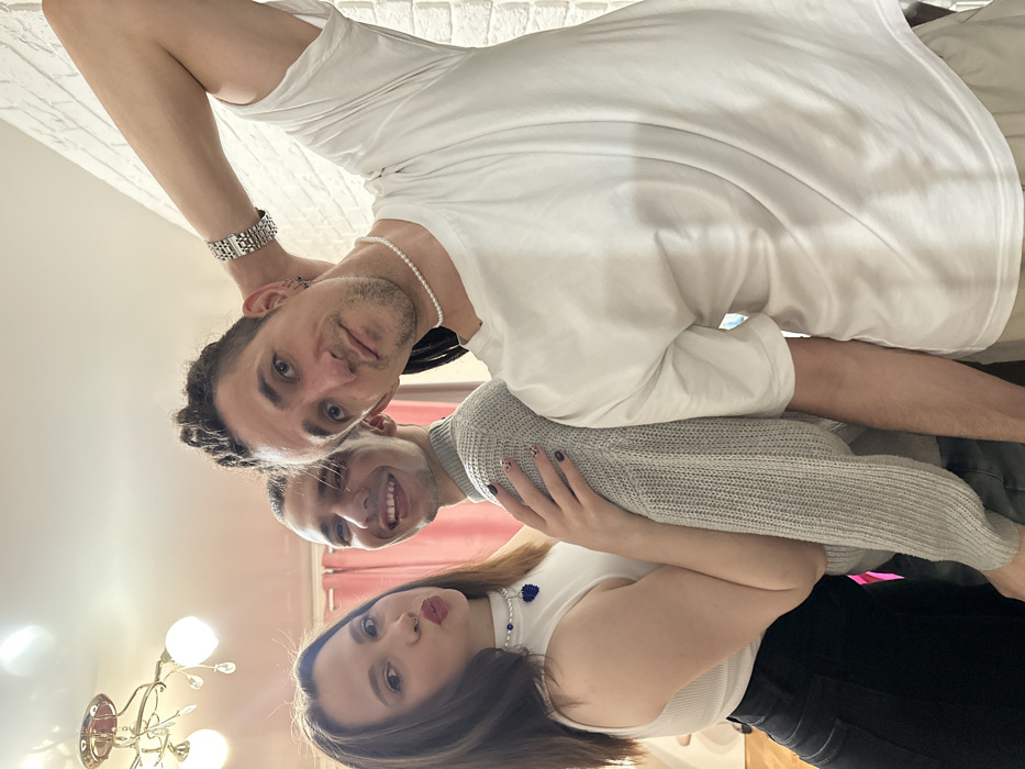 A group portrait features three individuals—one female and two males—standing in an indoor setting. From left to right: a female with dark, shoulder-length hair wears a white ribbed sleeveless top, black trousers, and a white pearl-like choker with a central blue, possibly beaded, pendant. She poses with pursed lips and her right hand resting on the mid-back of the adjacent male. The central male, with dark hair, wears a light grey ribbed long-sleeved sweater and smiles broadly, tongue visible. The male on the right, positioned slightly behind the central male, has dark dreadlocks, a white short-sleeved t-shirt, a white pearl-like choker, and a silver-colored watch on his left wrist. His left arm is bent with his hand placed behind his head. He looks directly at the camera with a neutral expression and has multiple small hoop earrings in his left ear. The background includes a light-colored ceiling with a multi-armed, gold-toned chandelier in the upper left, pink curtains to the left of the central male, and a white brick-patterned wall on the far right. No discernible landmarks or features are present within the image to confirm the location as Saratov, Russia, or any specific address within the city.