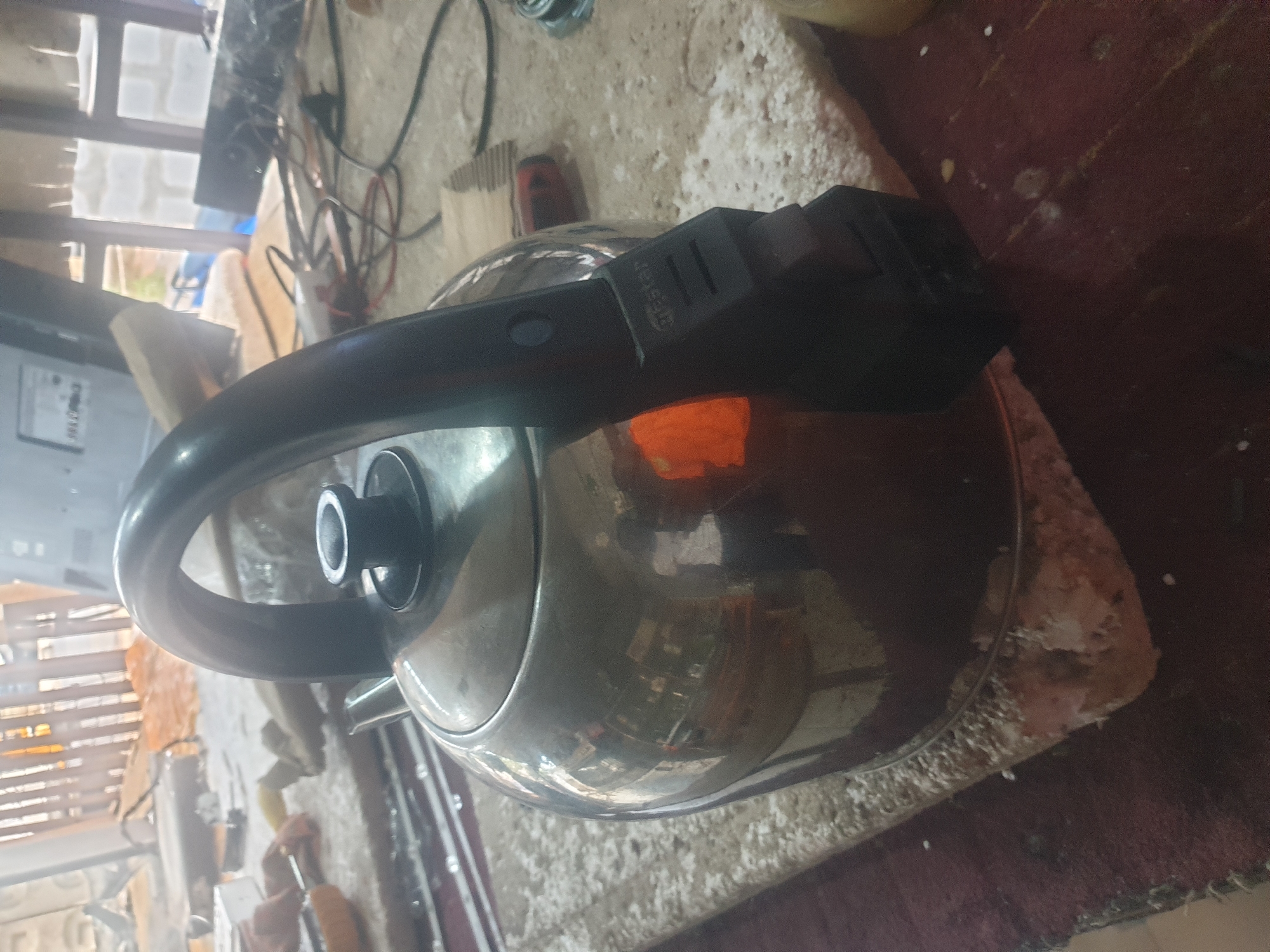 A close-up shot of a silver kettle with a black handle. The kettle appears to be sitting on a cluttered surface with a mix of items. In the background, there is a partially visible window or screen-like structure. The location of the scene is Unknown, Unknown.