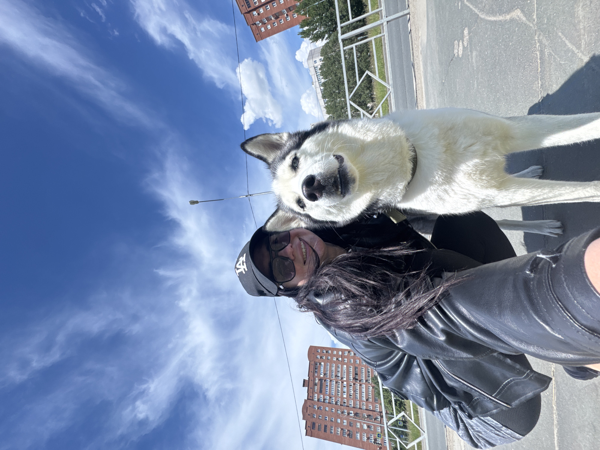 A selfie taken outdoors captures a woman and a dog against a bright blue sky with wispy clouds. The woman, wearing a black leather jacket, sunglasses, and a black baseball cap, appears to be holding the camera. Her dark hair flows down her shoulder. The dog, predominantly white with black markings around its eyes and ears, is positioned next to her. The scene appears to be an urban environment, with red brick buildings visible in the background. A metal railing runs horizontally across the frame behind the dog. The angle is tilted upwards, emphasizing the sky.