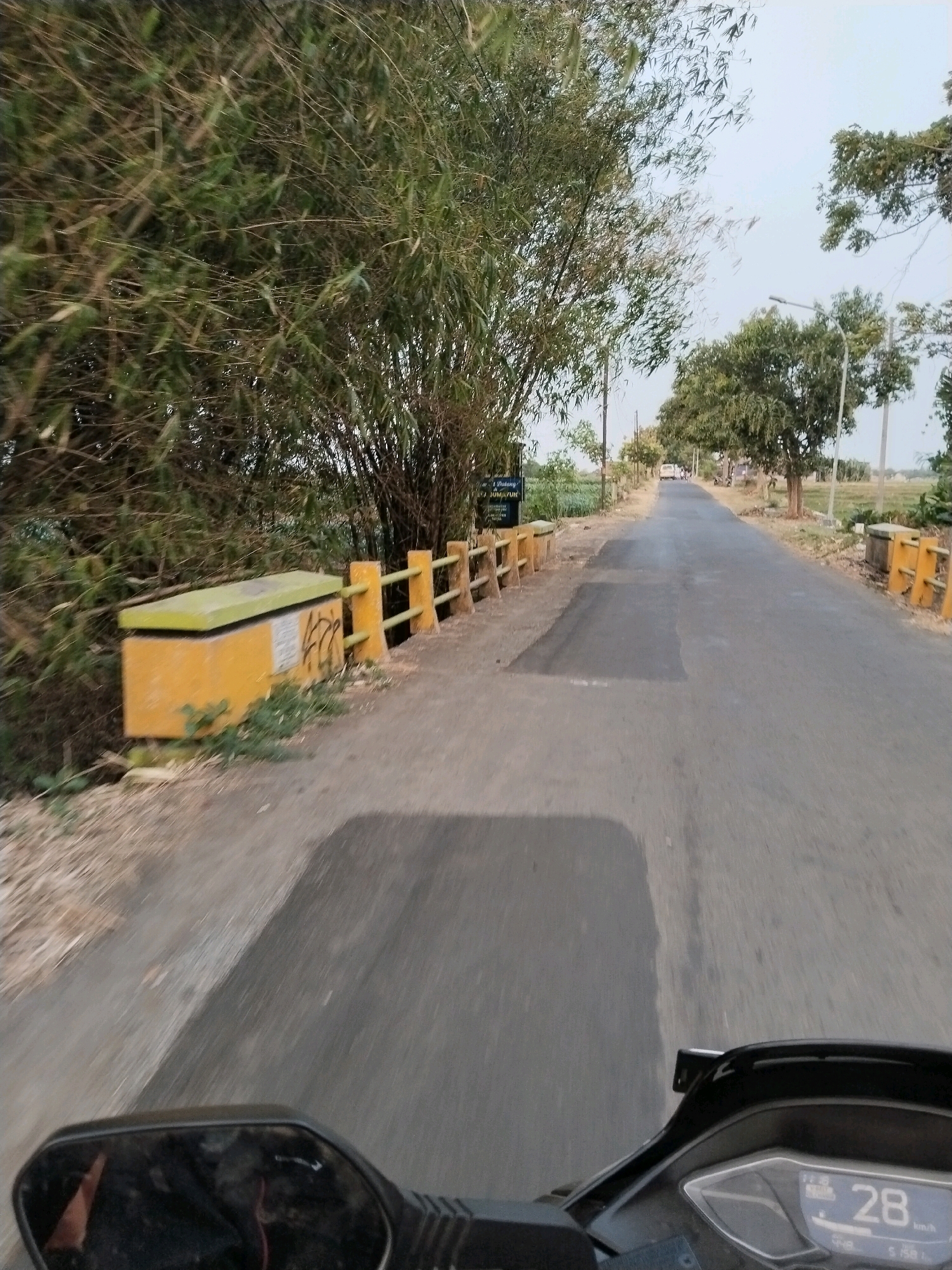 This image was captured from the perspective of someone riding a motorcycle on a road in Slarang Lor, Indonesia. The road is paved and appears to be a two-lane street with a slight curve to the right as it recedes into the distance. On the left side of the road, there is a dense thicket of bamboo plants and some dry grasses. A yellow, rectangular structure with a green lid is visible next to the bamboo, with graffiti on its side. A yellow railing with vertical posts borders the road in front of the bamboo. The right side of the road has more sparse trees and vegetation, with a paved walkway or shoulder. Further down the road, a few vehicles, including cars and what appear to be motorcycles, are visible. A street light is also present. In the foreground, the motorcycle's rearview mirror is visible, reflecting a person's arm. The motorcycle's digital dashboard displays a speed of 28 km/h. The sky is overcast and appears pale blue. The overall scene depicts a rural or semi-rural road in Indonesia during daytime.
