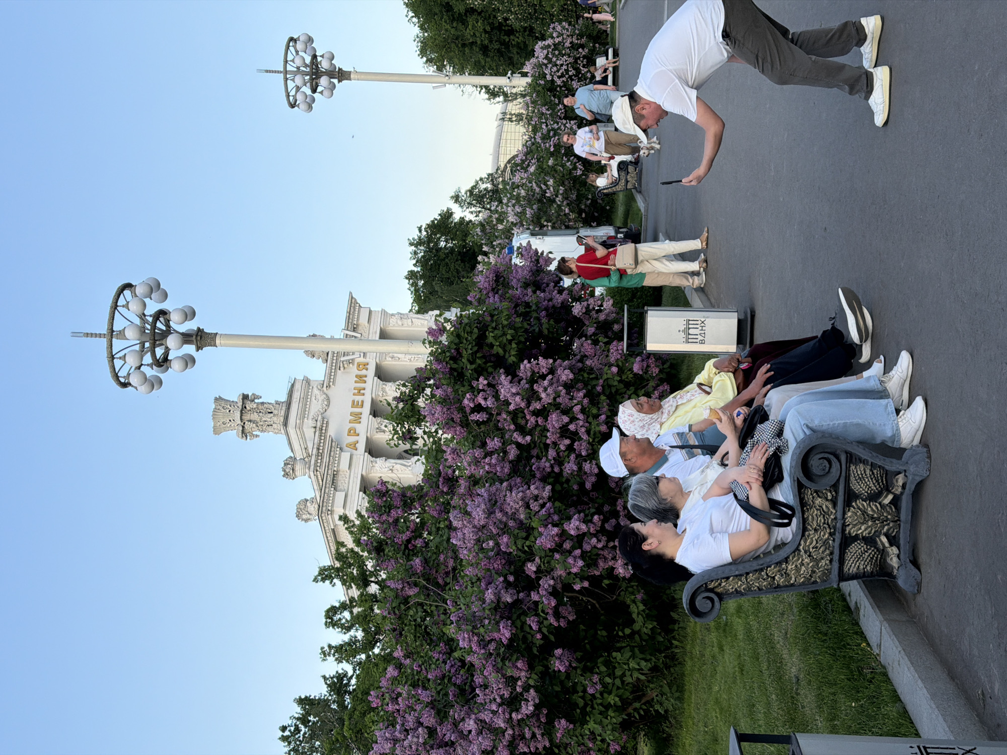 A medium, vertical shot captures a group of people in Moscow, Russia. The scene is set at the Exhibition of Achievements of National Economy (VDNKh), with the "Armenia" pavilion visible in the background. In the foreground, a group of four people are relaxing on a bench amidst blooming lilac bushes. The bench is black with gold details. A man in a white t-shirt and grey pants is bending over, apparently taking a photo with his phone. Other people are visible in the background, along with ornate lampposts. The sky is clear and blue.