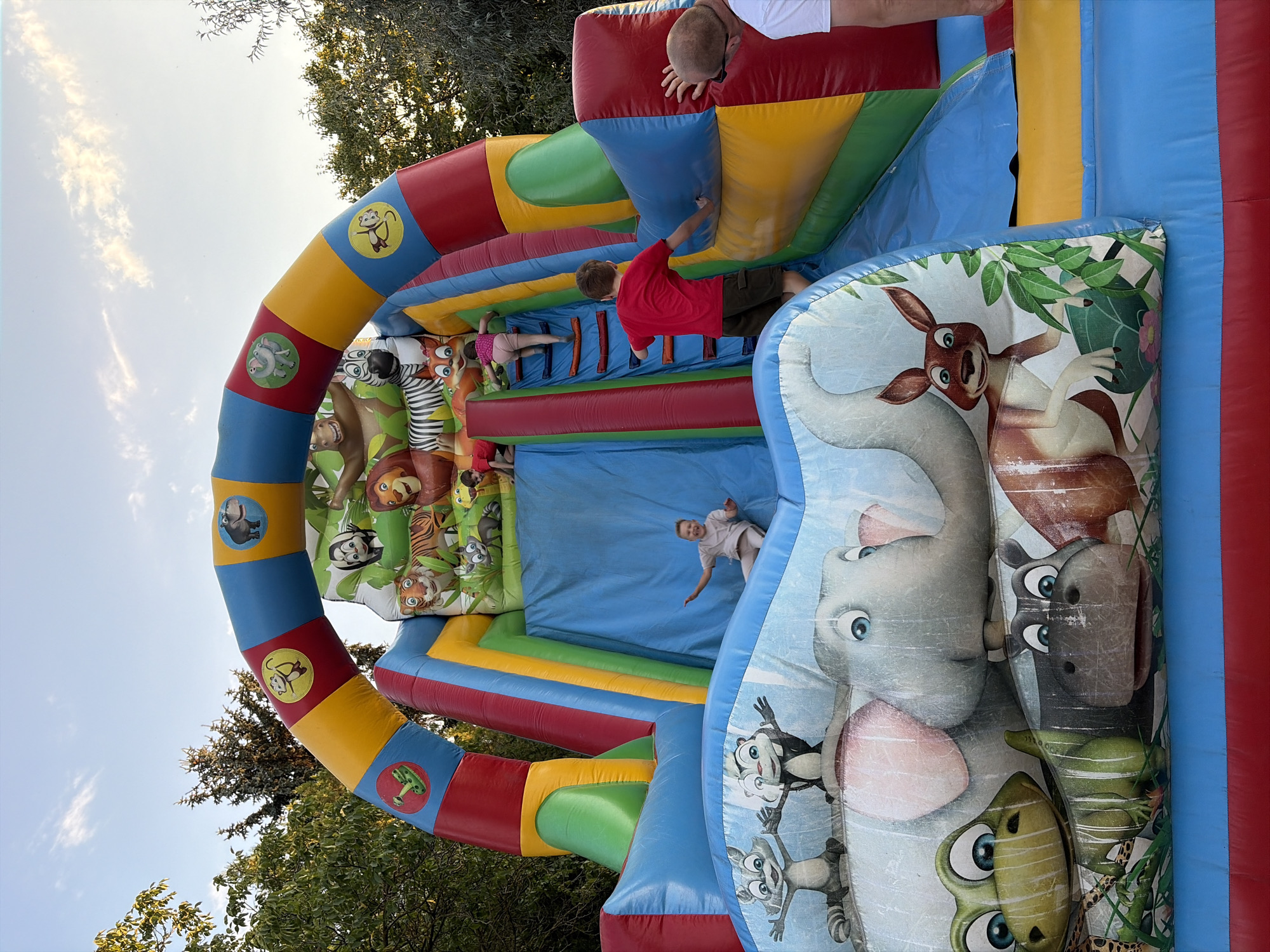The image shows an inflatable bouncy castle with a jungle theme, featuring cartoon animals such as an elephant, kangaroo, hippo, frog, raccoon, skunk, lion, tiger, and zebra. Several children are visible playing on the bouncy castle. One child is sliding down the blue slide, another is climbing the ladder on the right side of the slide, and a third child is also near the top of the slide area. An adult is partially visible on the far right, and another adult is partially visible on the left, facing away from the camera. The bouncy castle is set up outdoors, with trees and a blue sky with some clouds visible in the background. The location is identified as Oxentea, Moldova.