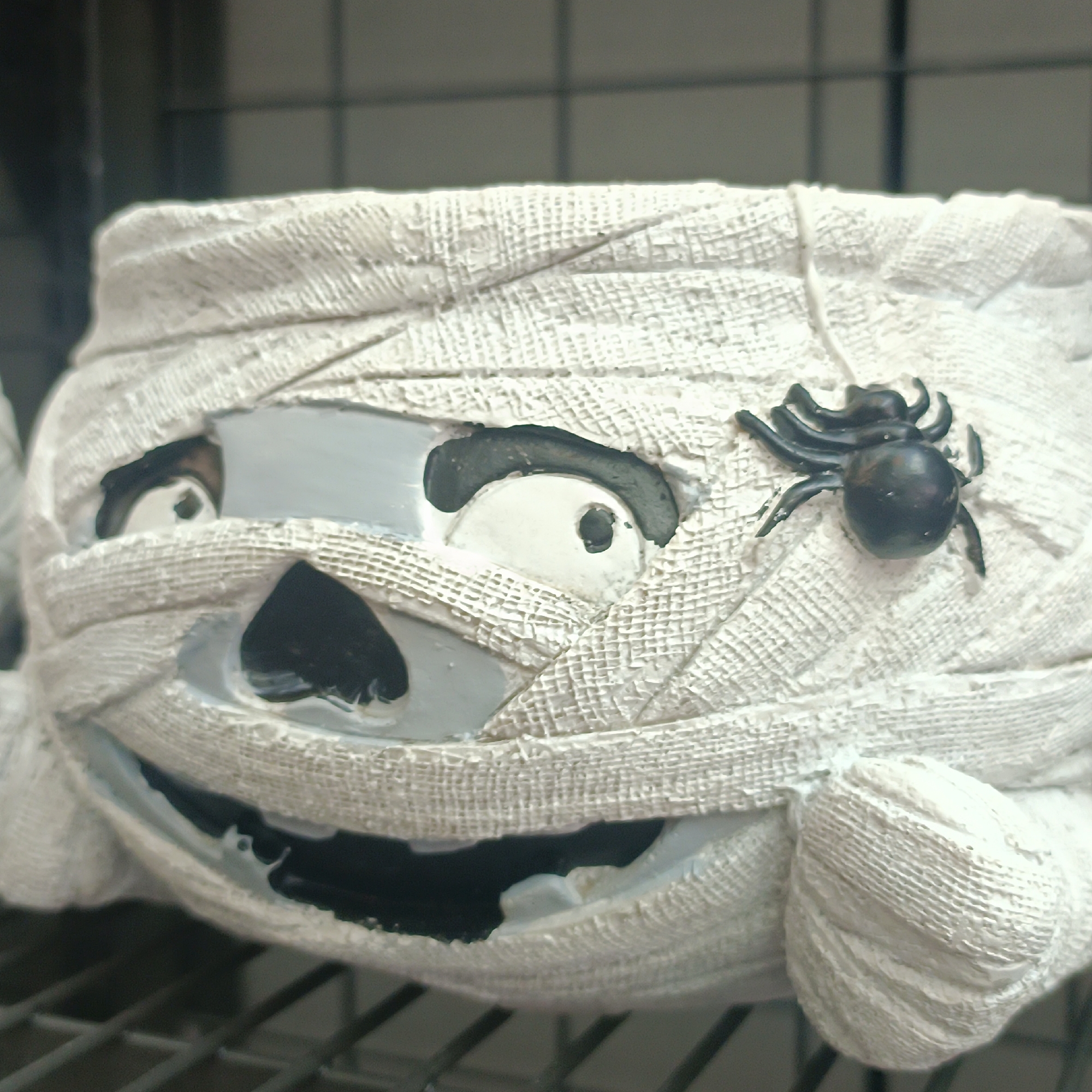 The image shows a close-up of a decorative Halloween planter shaped like a mummy. The mummy is white and appears to be made of a textured material resembling bandages. It has two large, round eyes with black pupils and a wide, dark mouth with a somewhat jagged opening. A small black spider decoration is positioned on the mummy's head. The planter is resting on a dark, slatted surface, possibly a shelf or rack, with a blurred background that suggests an indoor setting. There is no indication of the location being in Offenbach am Main, Germany.