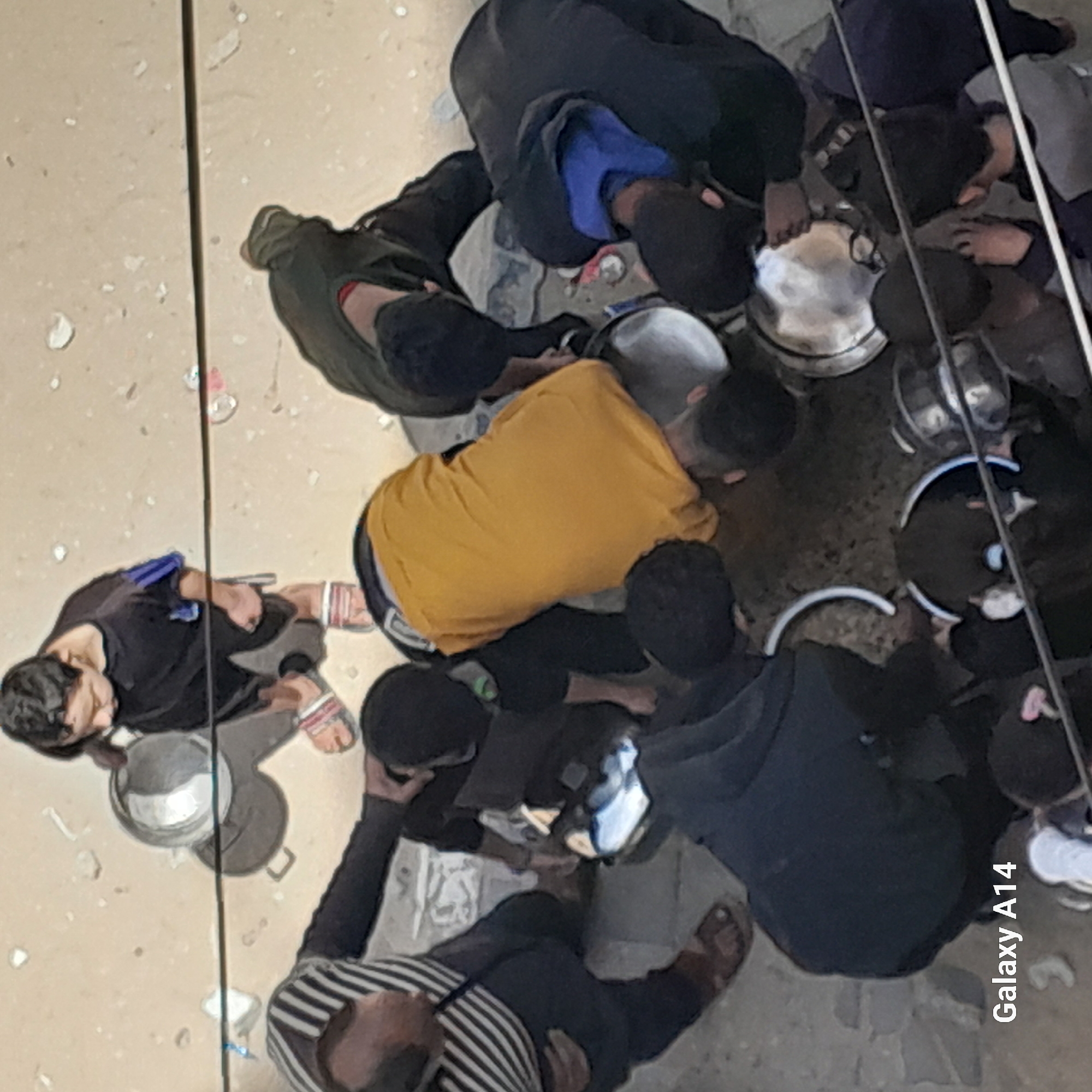 Several individuals are clustered together in an outdoor setting, possibly an alley or public space within a city. The scene appears to be taken from an elevated perspective. Most of the people are gathered around metal containers or pots. Many appear to be wearing dark or casual clothing. One person is wearing a bright yellow shirt. Footwear visible includes sandals and shoes. There are some white objects on the ground. The location is in the city of Unknown, Unknown.