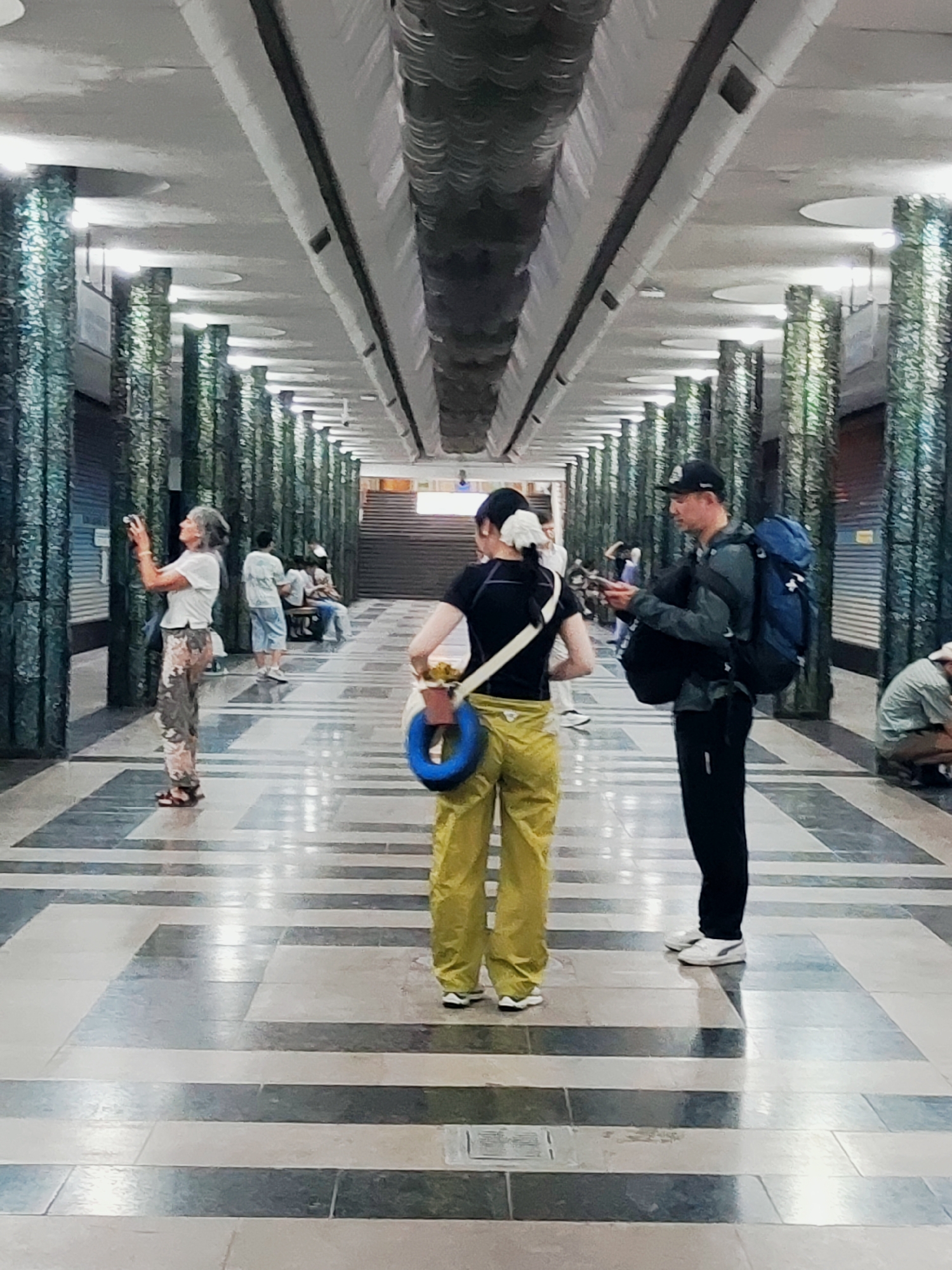 The image depicts a broad, covered walkway in Tashkent, Uzbekistan, likely a public space like a metro station or underpass. The walkway features a patterned floor with dark gray and lighter beige rectangular tiles arranged in a geometric design. On either side of the walkway are rows of tall, cylindrical pillars with a textured, greenish-blue mosaic or tiled surface. The ceiling is arched and illuminated by recessed lighting.
In the foreground, two individuals are the primary focus. A person wearing bright yellow pants and a black top is standing with their back to the camera, holding a blue ring and a small bag with a yellow strap. To their right, a man wearing a dark gray jacket, black pants, and a black baseball cap is looking down at his phone, holding a blue backpack on his left shoulder and a black duffel bag in his right arm.
Further in the background, other people are visible. On the left side, a woman with gray hair and patterned pants is holding a camera and looking up and to her left. Several other individuals are seated or standing further down the walkway. To the right, in the background, a person is kneeling. The overall atmosphere appears casual and public, with people engaged in various activities such as sightseeing, using their phones, or resting.