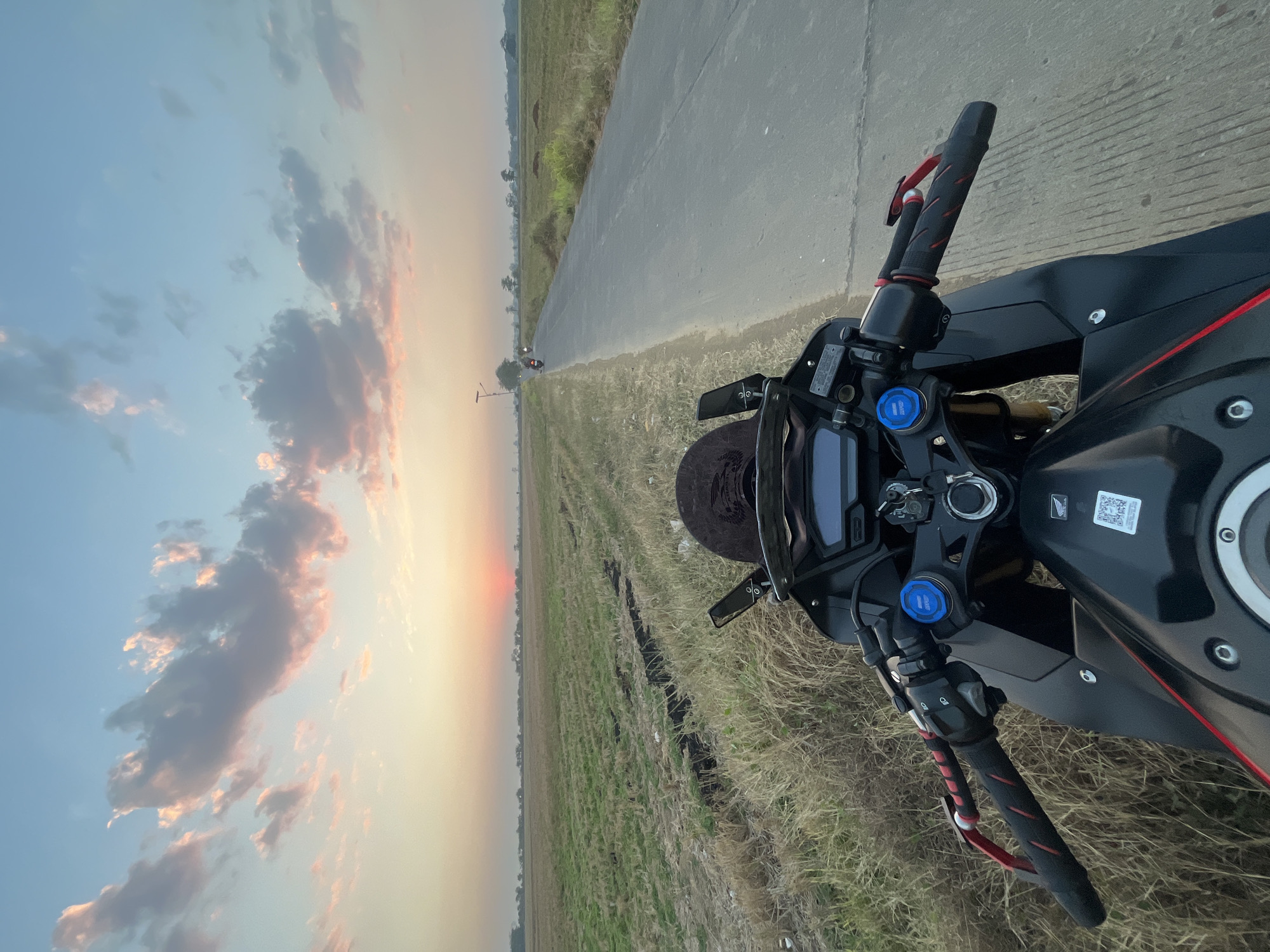 The image shows a first-person perspective of a person on a black motorcycle parked on the side of a rural road in Pecangaan, Indonesia, during sunset. The road stretches into the distance, lined with dry grass and a harvested rice field. In the background, a few scattered trees and a single lamppost are visible. The sky is a gradient of soft blues and oranges, with scattered clouds tinged pink and yellow by the setting sun. The motorcycle's handlebars, instrument panel, and front fairing are in the foreground. The scene evokes a sense of tranquility and the beauty of a rural sunset.