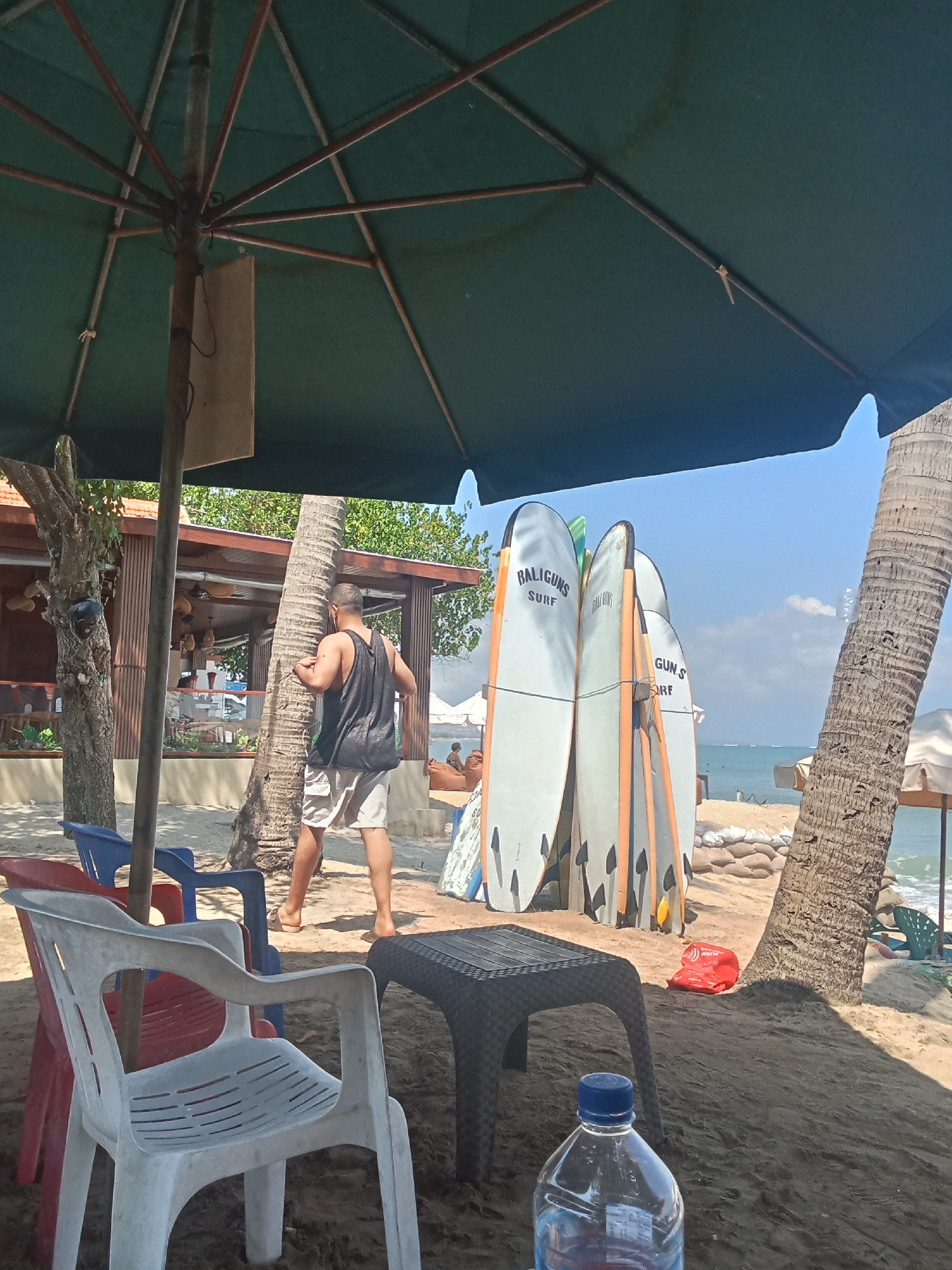 This image depicts a beach scene in Kuta, Indonesia. In the foreground, under a large green umbrella, are several plastic chairs, a black plastic table, and a partially visible bottle of water. To the right of the table are several surfboards leaning against each other. A man wearing a black tank top and light-colored shorts stands near a palm tree, facing away from the camera. He appears to be interacting with the tree. In the background, a beachside structure with wooden accents and open sides is visible, along with other beach umbrellas, sandbags, and more palm trees. The ocean and a clear blue sky are also present. The lighting suggests it is daytime.