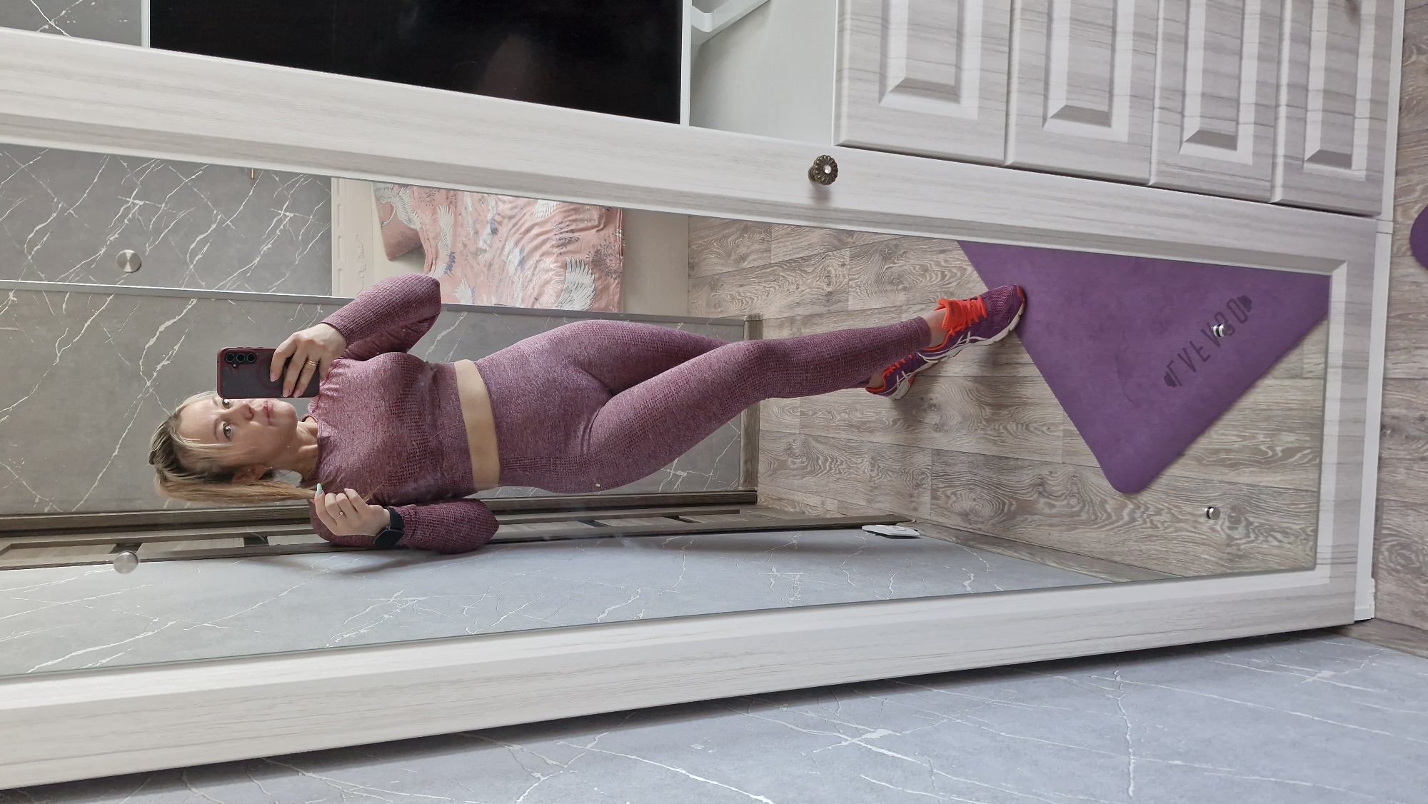 A mirror selfie captures a woman indoors. She is dressed in a matching workout set: a long-sleeved crop top and leggings, both in a textured reddish-purple hue. She wears sneakers with purple, white, and orange accents. Her blond hair is pulled back loosely. She holds a smartphone to take the photo. The background reflects parts of a room with gray and white walls. A yoga mat is visible, and glimpses of furniture and decor appear in the mirrored reflection.