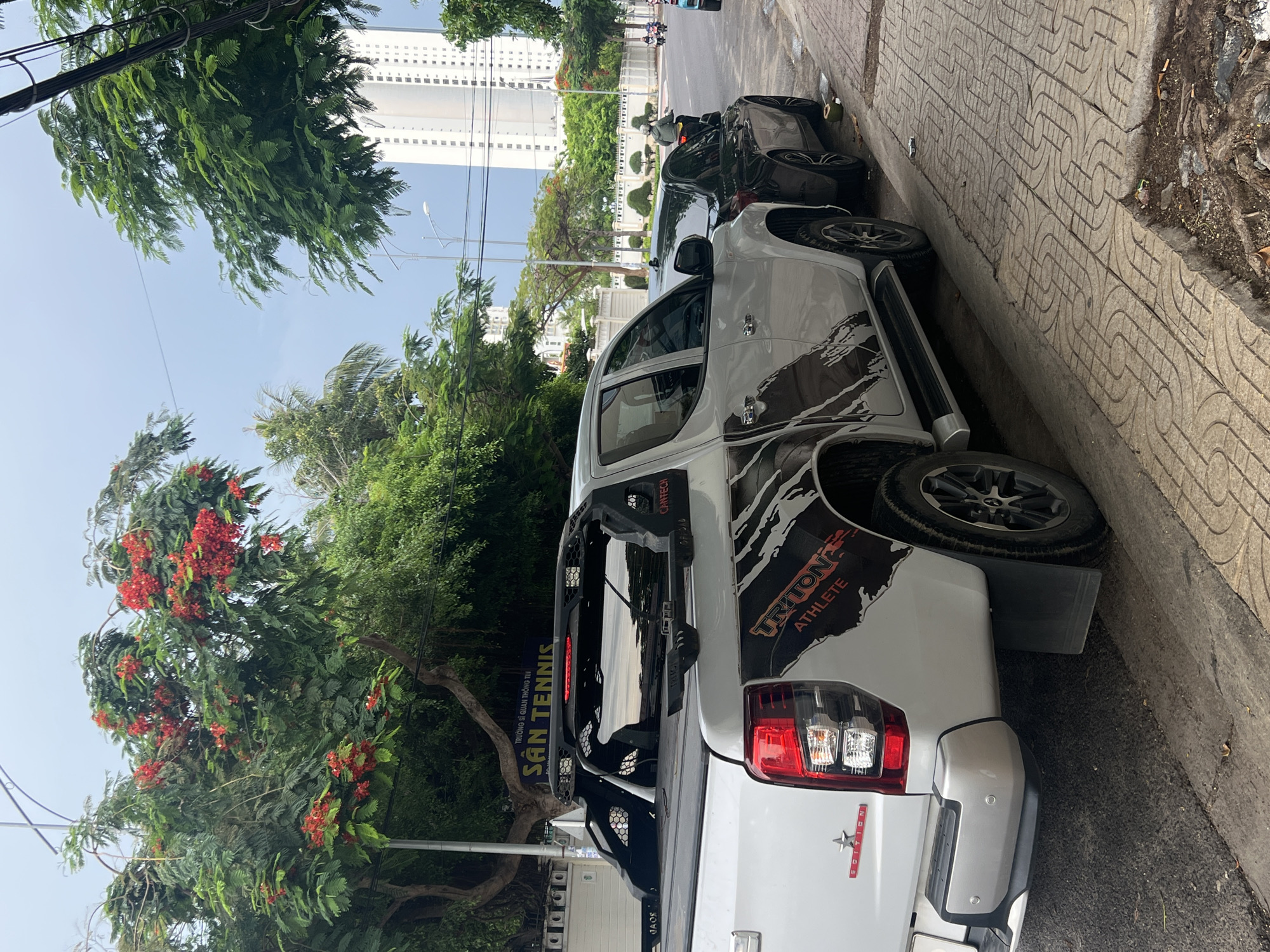 The image depicts a white Mitsubishi Triton Athlete pickup truck parked on a paved sidewalk in Bac Nha Trang Ward, Vietnam. The truck has black decals and aftermarket roll bars. Behind it, a dark-colored scooter or motorcycle is also parked. In the background, a tall building stands out in the urban landscape, with dense green trees alongside the street and red flowers in a tree branch. People are seen riding the scooter in the background.
