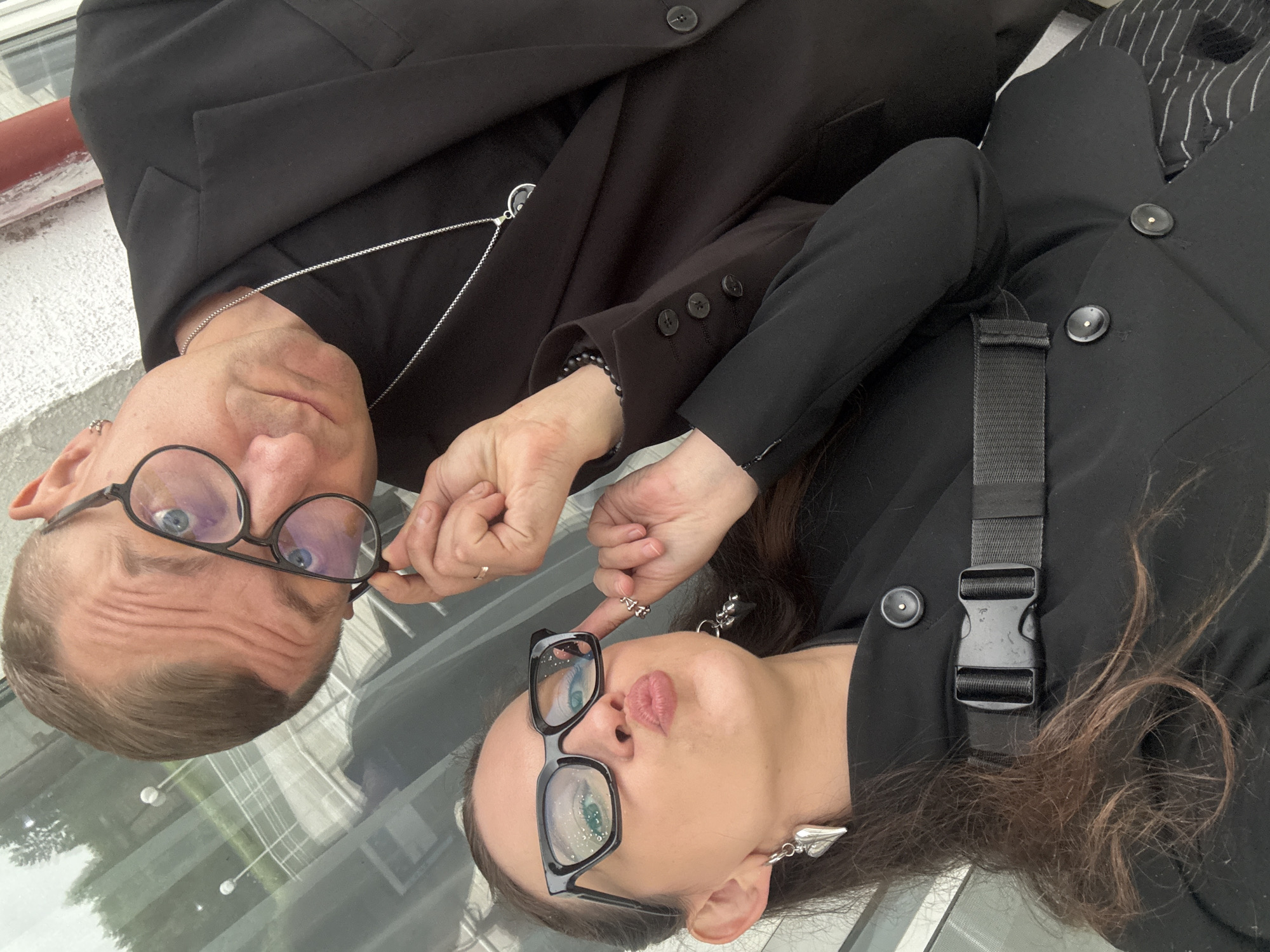 The image shows two individuals, a man and a woman, lying down and looking at the camera. They are both wearing black blazers and appear to be in a playful mood, with the woman poking the man's glasses and the man holding her finger. The man has short brown hair, is wearing glasses with black frames, and a silver chain necklace. The woman has long brown hair and is wearing large, cat-eye glasses with black frames and heart-shaped earrings.
The setting appears to be outdoors, possibly on a glass surface like a table or balcony, as indicated by the reflection of the sky and greenery below. The lighting suggests it is daytime. The overall mood is informal and intimate. No visible text is present in the image.