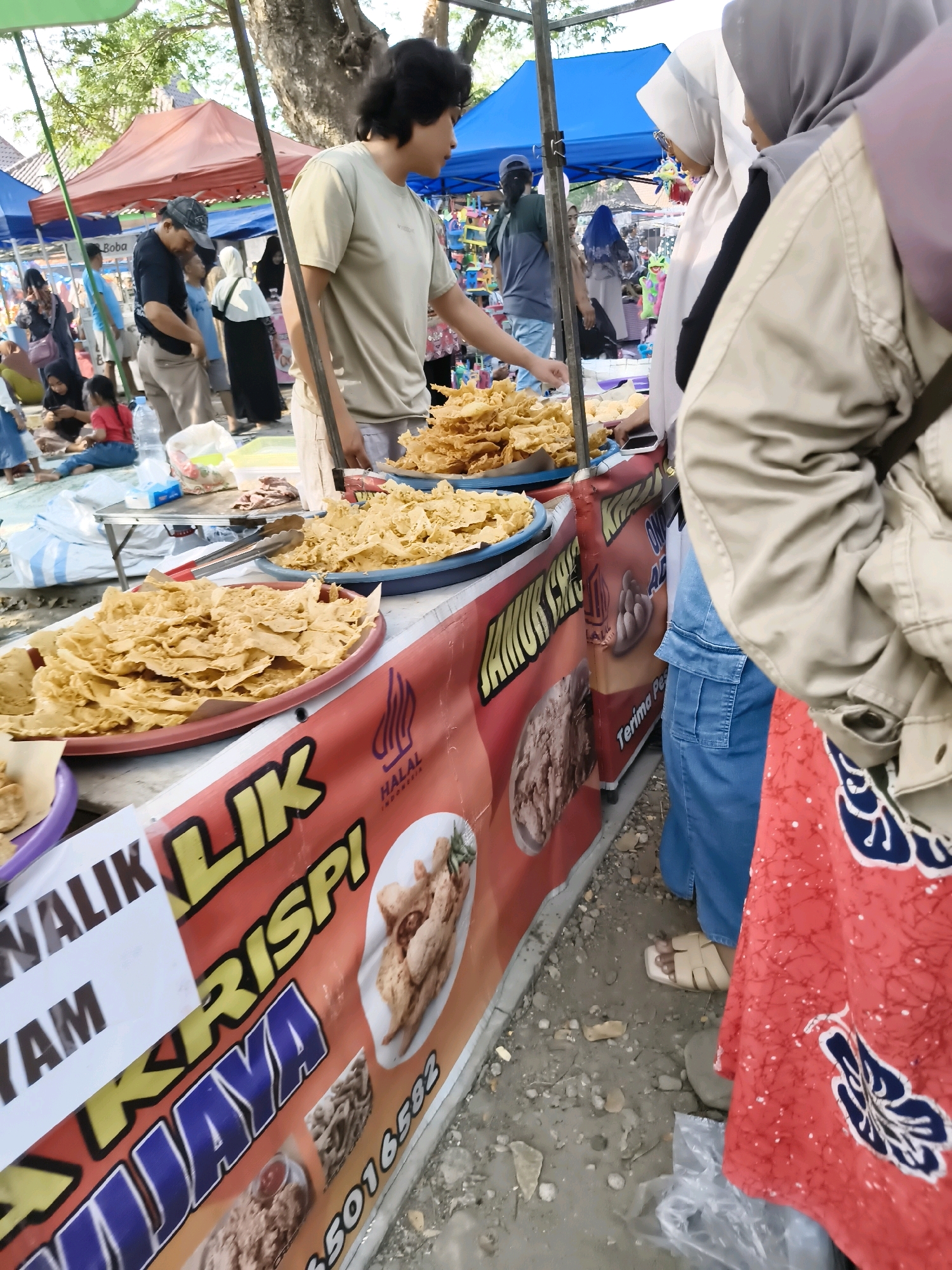 An outdoor market scene in Tlogorejo, Indonesia, features a food stall displaying crispy fried snacks. A vendor, a young man with dark hair, stands behind the stall, interacting with customers. To his right, two women wearing hijabs are browsing the snacks, with one woman in a light brown jacket and the other in a blue denim outfit. In the background, other people are visible, as well as tents and trees. The stall has a banner with Indonesian text and images of fried chicken and other food items. The ground is a mix of dirt and gravel, with some scattered debris.