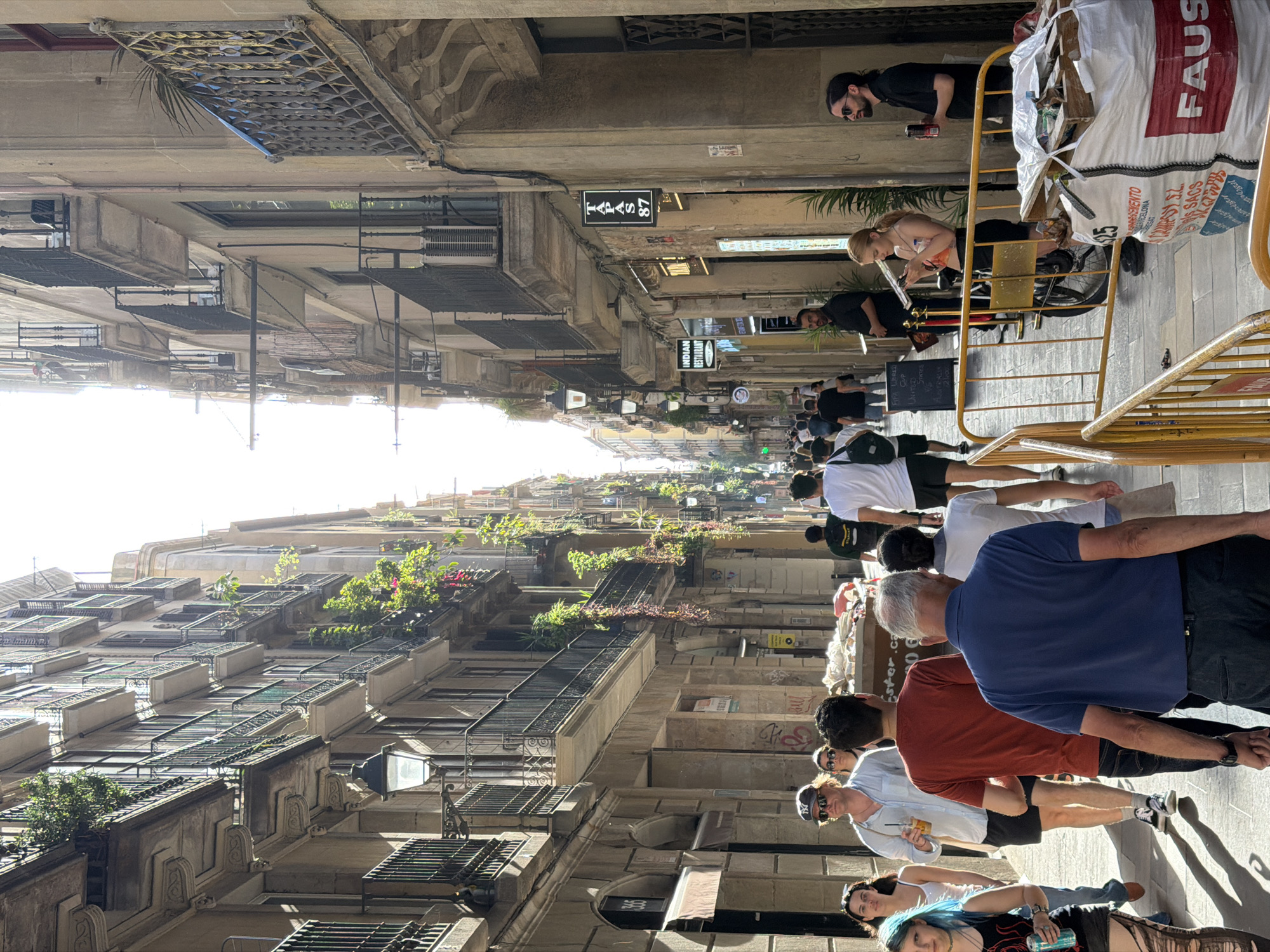 The image shows a narrow street in Barcelona, Spain, lined with tall buildings on either side. Sunlight streams down the street, creating strong shadows. Several people are walking in both directions. On the left, a woman with blue hair is visible, holding a can. Next to her, a man in a light-colored shirt and sunglasses holds a drink. Further down the street, a man in a blue shirt walks away from the camera. On the right side of the street, a sign reads "TAPAS 87". A woman is seated on a bicycle in front of a building, looking at her phone. A large white bag with red lettering is visible in the foreground on the right, with some wooden crates and debris on top of it. The buildings have balconies with railings, and some have plants and greenery. The street surface appears to be paved.