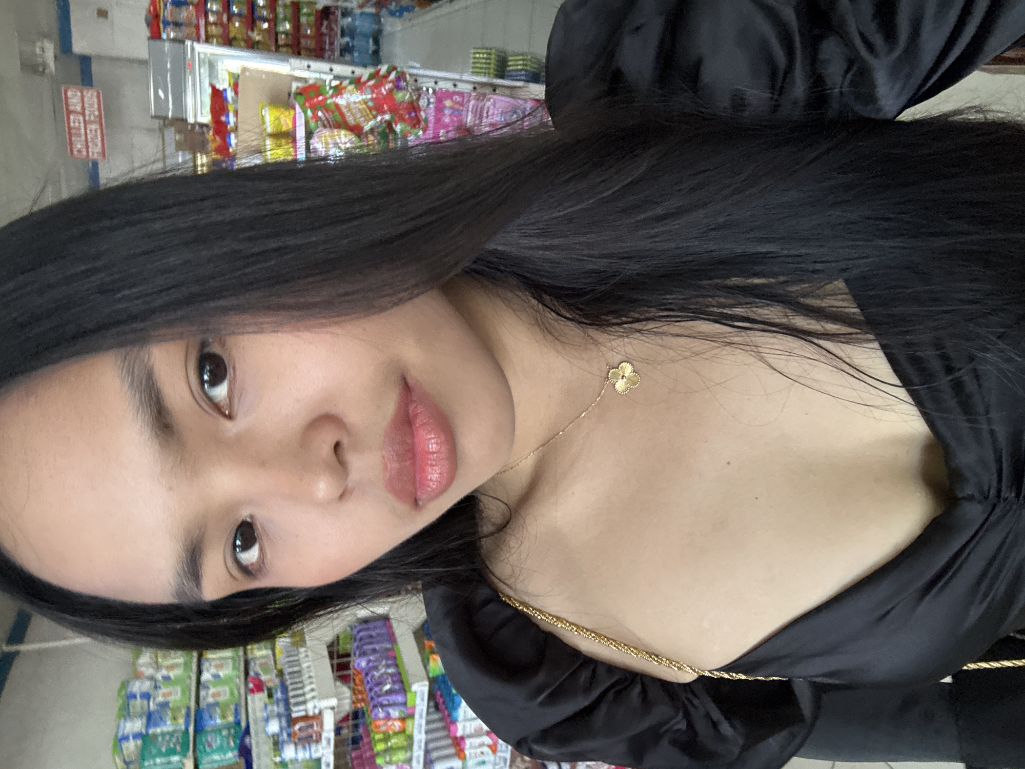 The image displays a selfie of a young woman taken inside a retail store, likely a convenience store or minimart in Davao City, Philippines. The photo is vertically oriented, with the woman's head turned towards the left of the frame. She looks directly at the camera with a calm, neutral expression.
The woman has long, straight, dark hair cascading over her shoulders. She wears a black top or dress with a gathered or ruffled neckline and sleeves. Adorning her neck are two gold necklaces: one featuring a prominent four-leaf clover pendant and another thinner chain. Her facial features include dark eyes and full, slightly parted lips.
The background is an out-of-focus retail environment, showing numerous shelves stocked with various packaged items, including snacks and beverages. A prominent red rectangular sign is visible on the left side of the background, rotated, and clearly displays the text "CHILLED AND FROZEN FOODS." The indoor setting is illuminated by artificial lighting, suggesting the photo could have been taken at any time of day. No specific activity is depicted beyond the woman taking a selfie.
