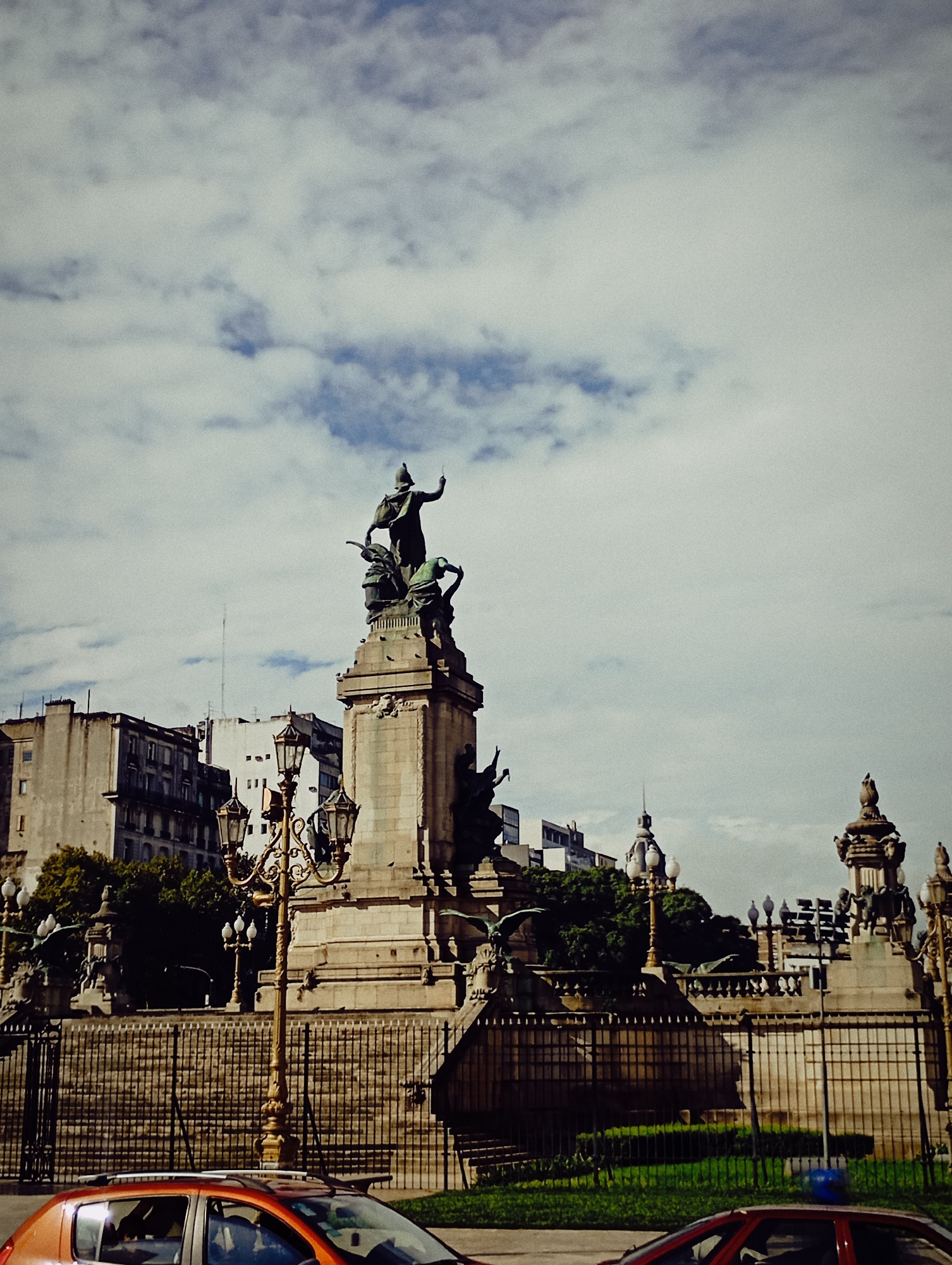 The image displays a grand public monument in an urban setting, likely Buenos Aires, Argentina, under an overcast sky. The central subject is a tall, ornate stone monument crowned with bronze sculptures. The most prominent bronze figure stands on a high pedestal, depicted as a classical or allegorical male figure with an arm raised, wearing a helmet or cap and a flowing garment. Below this main statue, other smaller bronze figures or decorative elements are integrated into the monument's base.
The monument is enclosed by a dark, decorative metal fence with pointed finials, separating it from the street. An elaborate, golden-colored street lamp with multiple glass globes and intricate scrollwork stands to the left of the monument. In the background, several multi-story buildings with varied architectural styles, typical of an established city, line the streets. Some greenery, possibly grass or bushes, is visible behind the fence.
In the foreground, the upper parts of two cars are visible: the roof and windshield of an orange-red car on the left, and a portion of a red car on the right. The sky is largely covered with light grey and white clouds, indicating an overcast daytime. The scene is static, with no discernible human activity. No visible text is legible in the image.