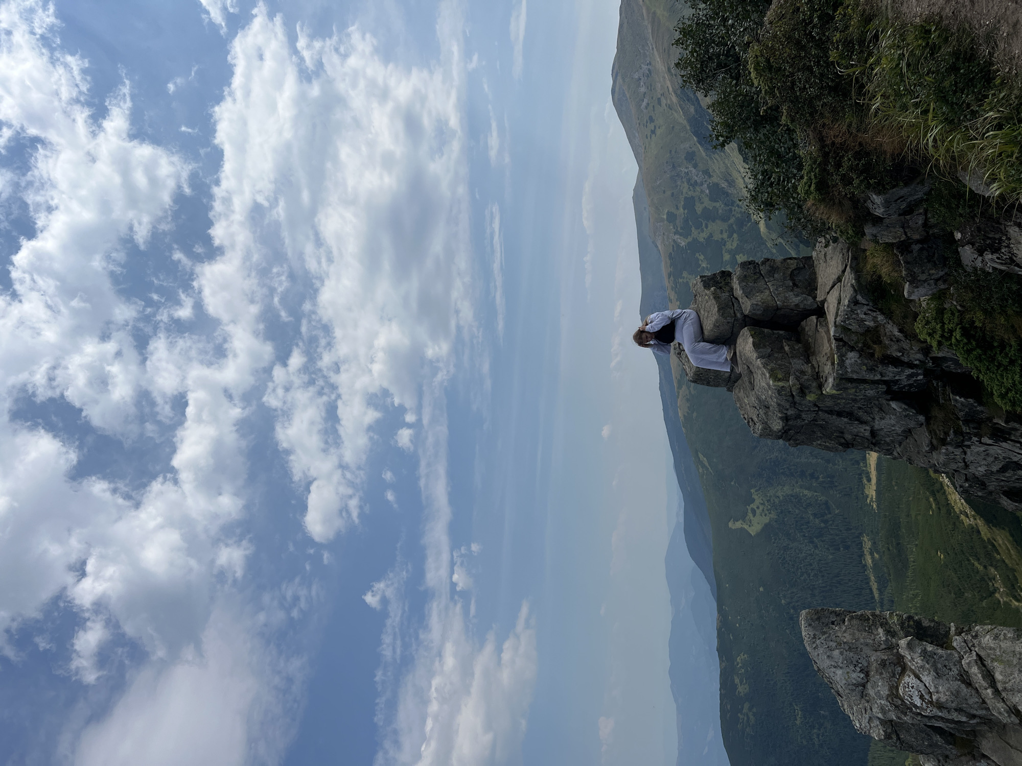 A person is sitting on a rocky outcrop in the Carpathian Mountains, near Vorokhta, Ukraine. The person is wearing white athletic wear and has their hands on their head. The background shows a vast mountainous landscape with rolling hills covered in green vegetation and forests. The sky is blue with scattered white clouds. In the foreground, large rock formations frame the scene.