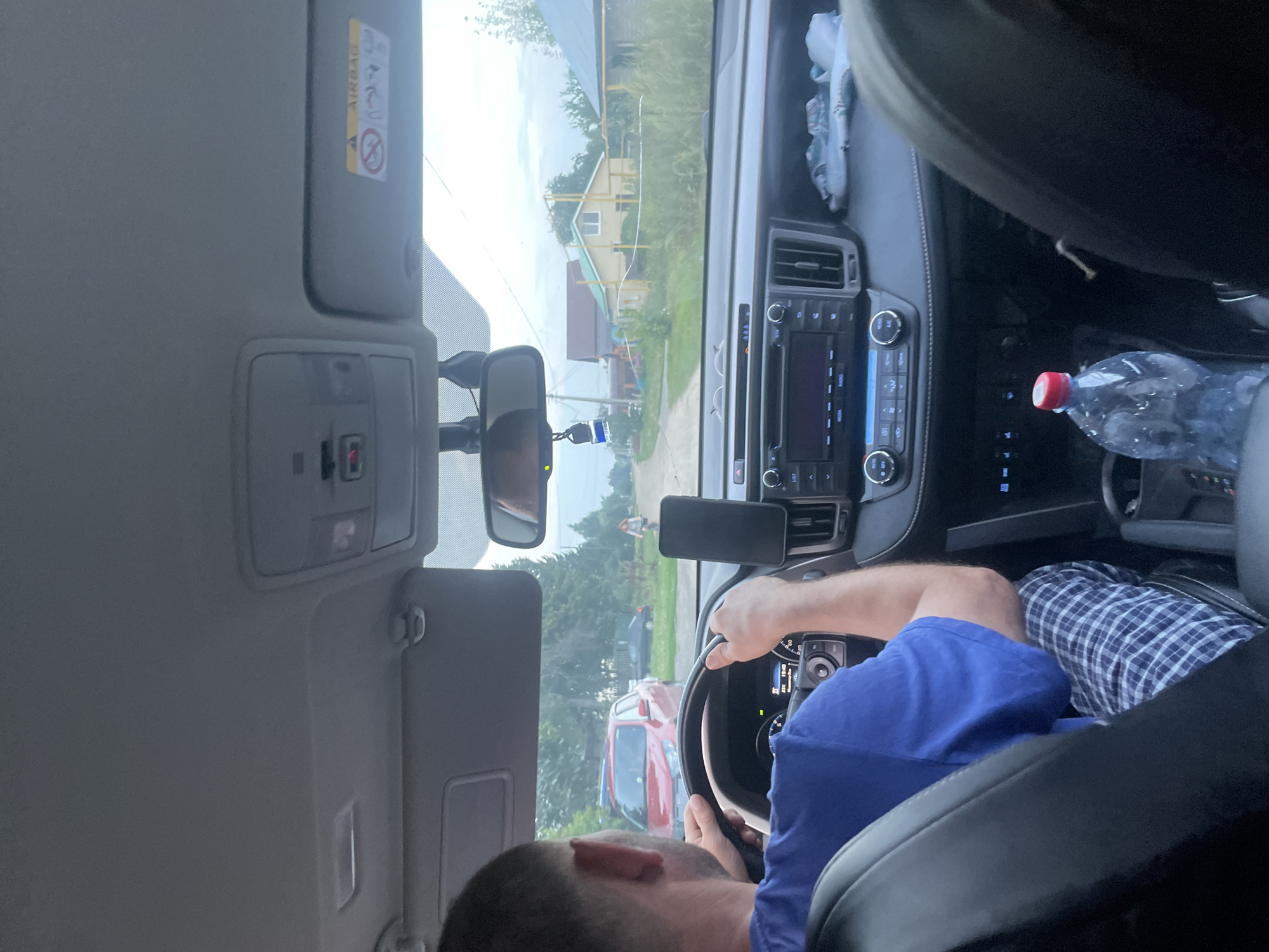 The image shows the interior of a car from the perspective of the passenger seat, looking forward. A person wearing a blue t-shirt and plaid pajama pants is driving, their hands on the steering wheel. The dashboard displays "25 C 19:40". Outside the car, a dirt road is visible with houses and trees lining it. A red car is parked on the side of the road. A person is riding a bicycle in the distance. A plastic bottle of water is on the center console. A yellow gas pipe is visible in front of a yellow house. The image appears to have been taken in a rural area, consistent with Laishevo, Russia, based on the architecture and vegetation.
