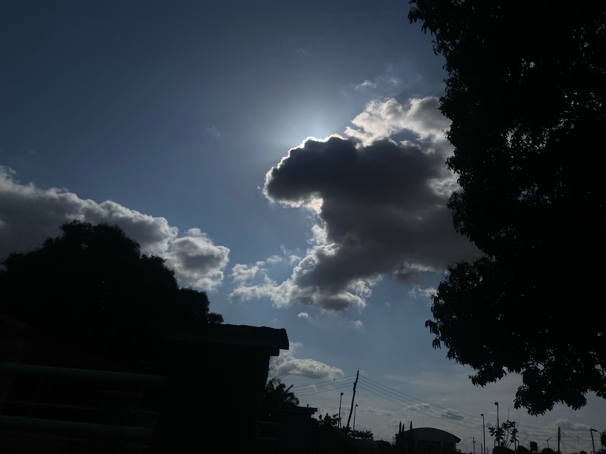 The image shows a cloudy sky with the sun partially obscured by a large, dark cloud. The sun's rays are visible, creating a bright halo effect. In the foreground, silhouetted against the sky, are trees and buildings. A portion of a building with a dark roof is visible on the left, and a large tree with dense foliage is on the right. Utility poles and wires are also present in the background, suggesting an urban or suburban setting. The overall impression is a dramatic sky scene, likely captured during daylight hours in Barden Yana, Nigeria.