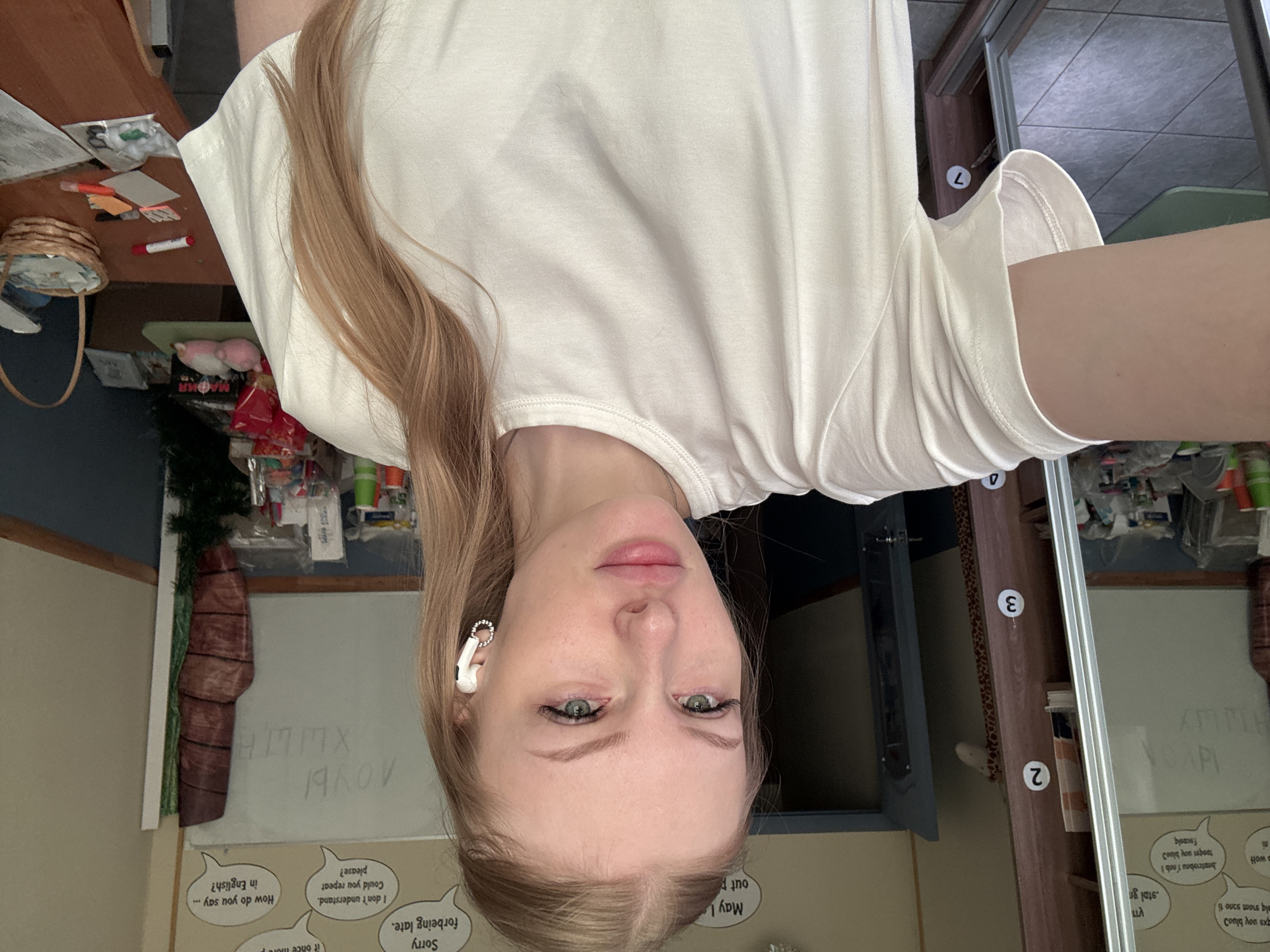 A young woman with blonde hair and green eyes is taking a selfie in what appears to be a classroom or study area. She is wearing a plain white t-shirt and white earbuds. Behind her, a whiteboard with writing is visible, along with a bookshelf and various classroom materials. The overall impression is casual and informal, possibly a personal space for learning or work. The presence of numbered stickers on a shelf suggest a possible organizational system or a playful touch. The environment looks somewhat cluttered but functional, with items like books, supplies, and a basket on a table. The lighting suggests it's daytime, with natural light contributing to the overall illumination.