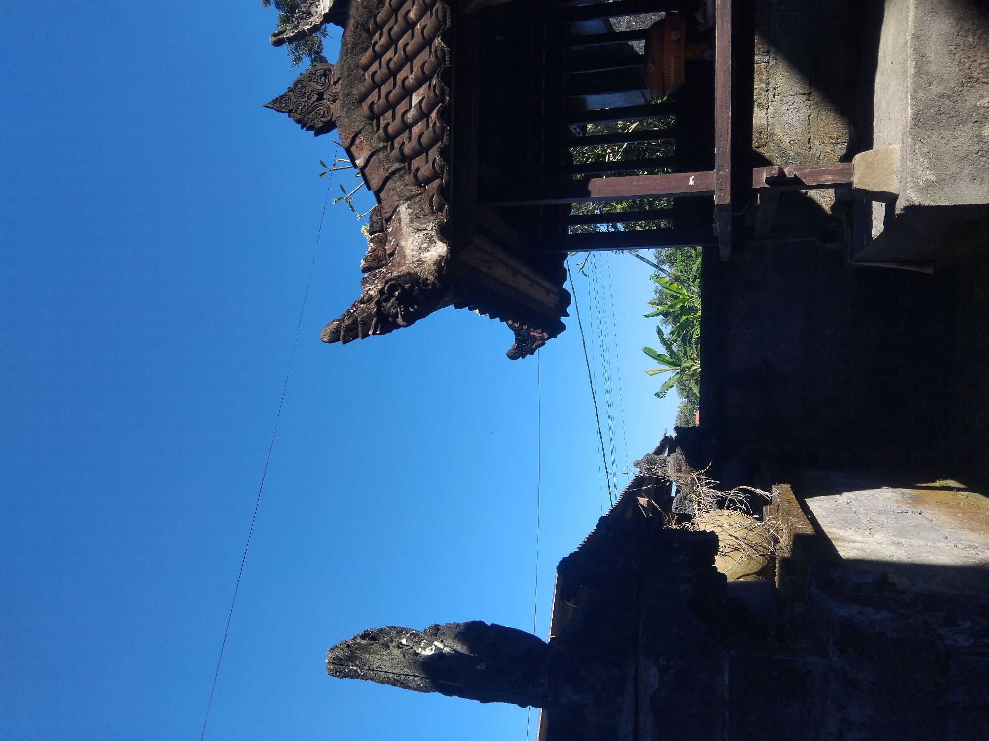 A low-angle shot depicts portions of a weathered structure, likely a building or temple, against a clear blue sky. The building features a traditional tiled roof with ornate carvings on its edges. In the background, vegetation including palm trees can be seen. The structure appears to be aged and possibly abandoned, with signs of weathering and discoloration. The photograph appears to be taken during daylight hours, with the sun illuminating certain parts of the building.